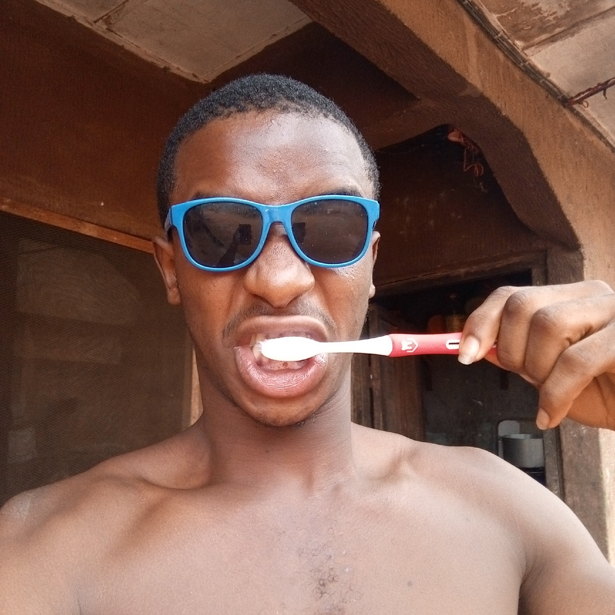 A dark-skinned, shirtless man with short hair is seen brushing his teeth outdoors. He wears bright blue sunglasses. He holds a red and white toothbrush in his right hand, and the toothbrush is in his mouth. The background suggests an outdoor or semi-outdoor setting, possibly a porch or veranda. The architecture visible in the background is simple and appears to be typical of a residential area. The location is Onitsha, Nigeria based on contextual information.