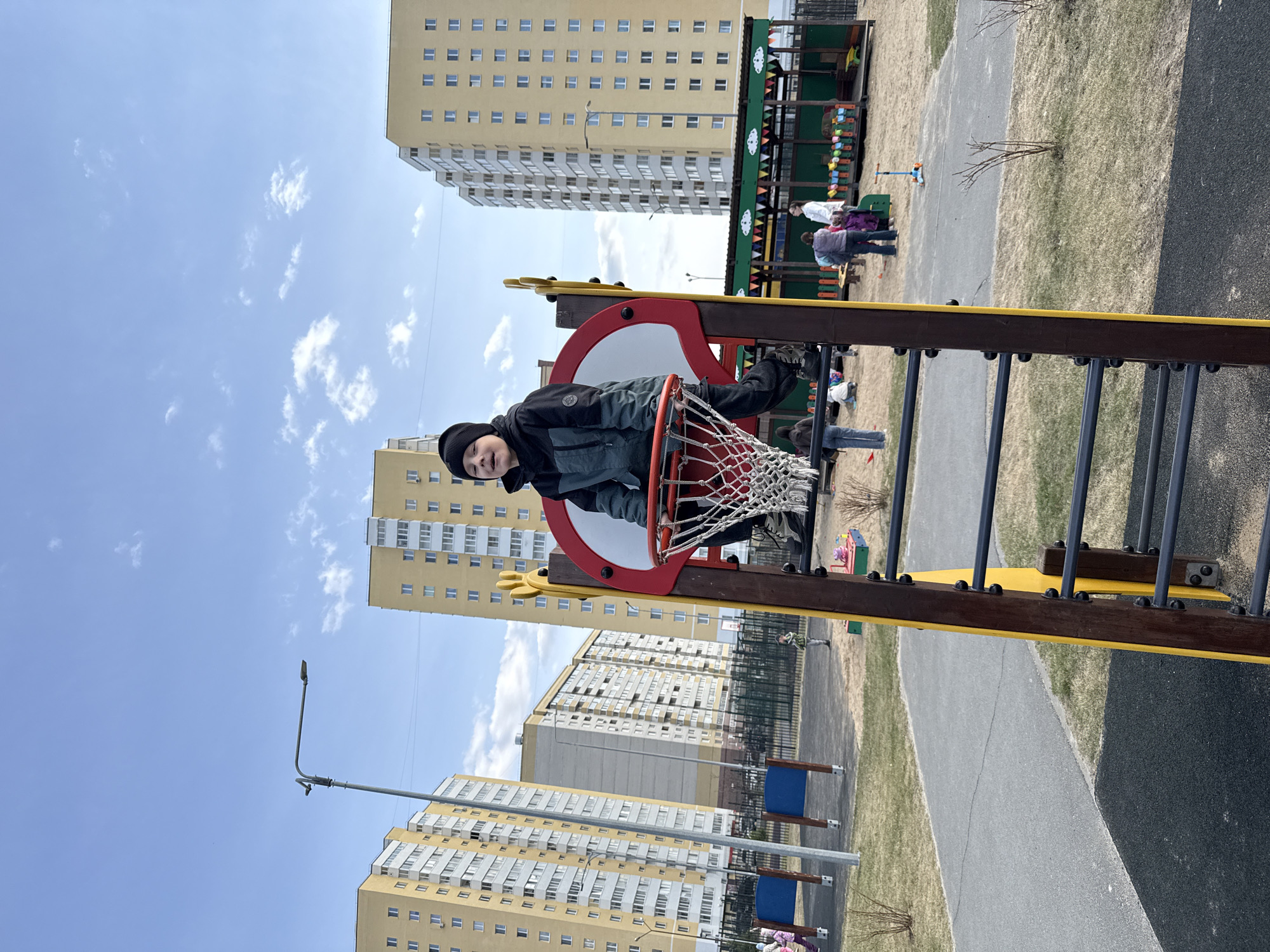 This image, taken in Tobolsk, Russia, captures a vibrant scene at a children's playground in an urban residential setting on a clear, sunny day.
The main subject is a young boy, positioned playfully inside a basketball hoop, which is part of a larger red, yellow, and brown playground structure. Dressed in a dark jacket, dark pants, and a black beanie, he looks directly at the camera with a subtle smile. His left leg is visible through the net of the hoop, suggesting he is climbing or sitting within it.
In the background, multiple tall, light-yellow apartment buildings with numerous windows dominate the skyline, indicative of a modern housing development. The bright blue sky, adorned with scattered white clouds, suggests pleasant weather. The ground areas consist of patchy grass, sandy sections, and paved pathways.
Several other people, including adults and children, are visible in the mid-to-far background. They appear to be enjoying the playground facilities or walking nearby, contributing to the lively atmosphere. One adult in a white coat and a child in purple clothing are noticeable on the right side of the background. A street lamp can also be seen against the sky. The image conveys a cheerful and active moment at a community playground. There is no discernible text visible within the frame.