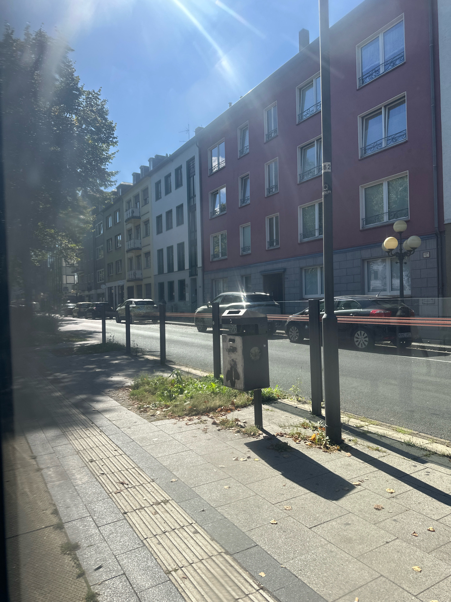 This image depicts a street scene in Krefeld, Germany, on a sunny day. The perspective is from a sidewalk looking across a street lined with multi-story buildings.
In the foreground, a textured, tactile paving strip runs along the edge of the sidewalk, indicating a pedestrian pathway. The sidewalk itself is paved with grey rectangular tiles. Sunlight casts long shadows across the sidewalk and onto the street.
The street features parked cars on the left side, and moving traffic on the right. Several cars are visible, including a dark colored station wagon with license plate KR-RC 1911. A large trash receptacle with a sticker is positioned on the sidewalk. Street lamps with multiple globes are visible along the street.
The buildings lining the street are varied in color and architectural style, with multiple windows, some featuring decorative ironwork. The sky is bright blue with visible sun glare. Trees with green foliage are present on the left side of the image, casting shadows. Scattered leaves are visible on the sidewalk and the strip of grass between the sidewalk and the street.
