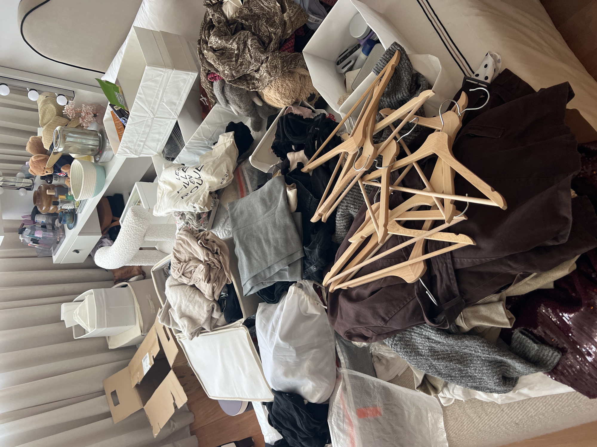 The image depicts a cluttered bedroom scene, likely within a residence, possibly in Porto, Portugal, given the context of the request. The primary focus is a bed laden with a disorganized collection of clothing and storage containers. Several fabric storage bins, some containing folded or bundled garments in neutral tones such as beige and light brown, are visible. A large cardboard box, partially opened, lies on the floor near the bed.
Dominating the foreground is a large pile of wooden clothes hangers, some still holding garments. Interspersed with the hangers are various articles of clothing, including sweaters in grey and brown, a dark brown jacket, and what appears to be sequined fabric. Other objects visible include a white laundry bag, a grey fabric item that could be a cushion or stuffed toy, and patterned fabrics resembling scarves or shawls.
In the background, a white dressing table with drawers is partially visible, adorned with makeup brushes in containers, bottles, and decorative items like a teddy bear and a straw hat. Behind the dressing table, light-colored curtains obscure the view of any window. The overall impression is one of disarray and a room undergoing a process of packing, unpacking, or general tidying. There are no people visible in the image.