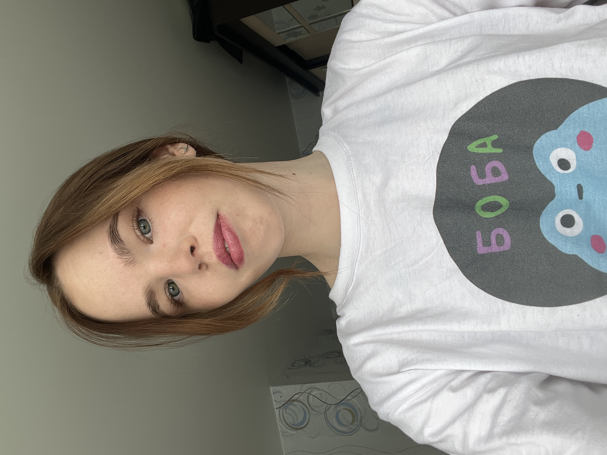 A fair-skinned person with shoulder-length auburn hair and light green eyes is facing the camera. They are wearing a white long-sleeved t-shirt with a black circle graphic on the right side, featuring a light blue cartoon character with pink and white details. The word "БOBA" (BOBA) is written in green and purple within the circle. The background consists of a light gray wall, and a dark, blurred object suggests furniture or a fixture in the upper portion of the frame. Below the person, a wall decorated with blue, white, and gray circles is visible. The scene appears to be indoors and is not visually consistent with outdoor locations in the city of Chaikovsky, Russia, based on typical architectural and environmental features.