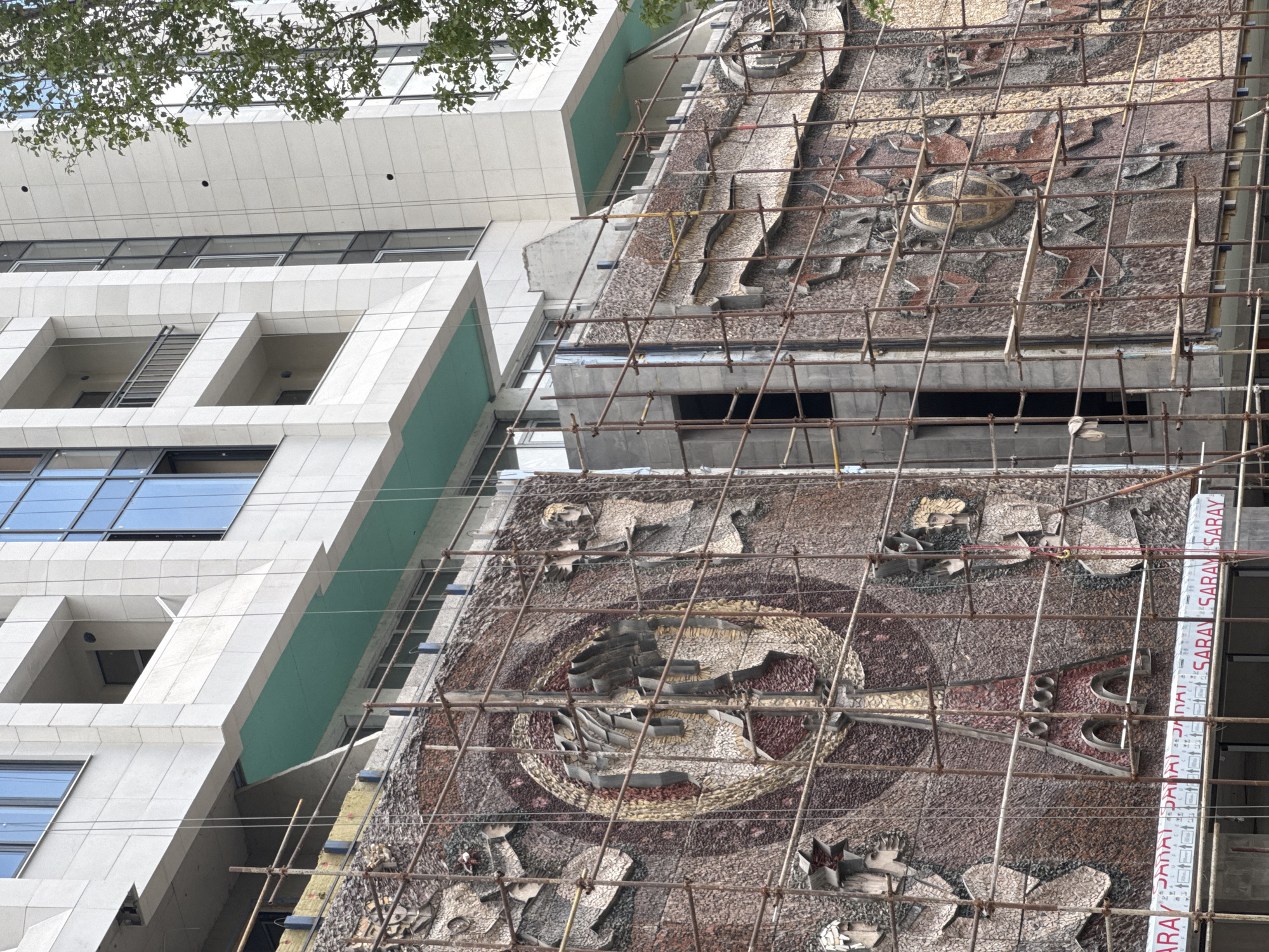 The image depicts a partial view of a building undergoing renovation in an unknown city. A section of the building's facade is covered with a large, mosaic artwork, which features figures and abstract designs in various shades of brown, grey, and reddish hues. The mosaic art appears to be under restoration, as scaffolding is erected in front of it. A modern, white building with large windows and green accents is visible next to the mosaic-covered structure. A ladder is positioned against the side of the white building. The scaffolding features several white panels with the text "SARAY" printed on them. A few tree branches with green leaves are visible in the upper left corner of the image, suggesting an outdoor setting.
