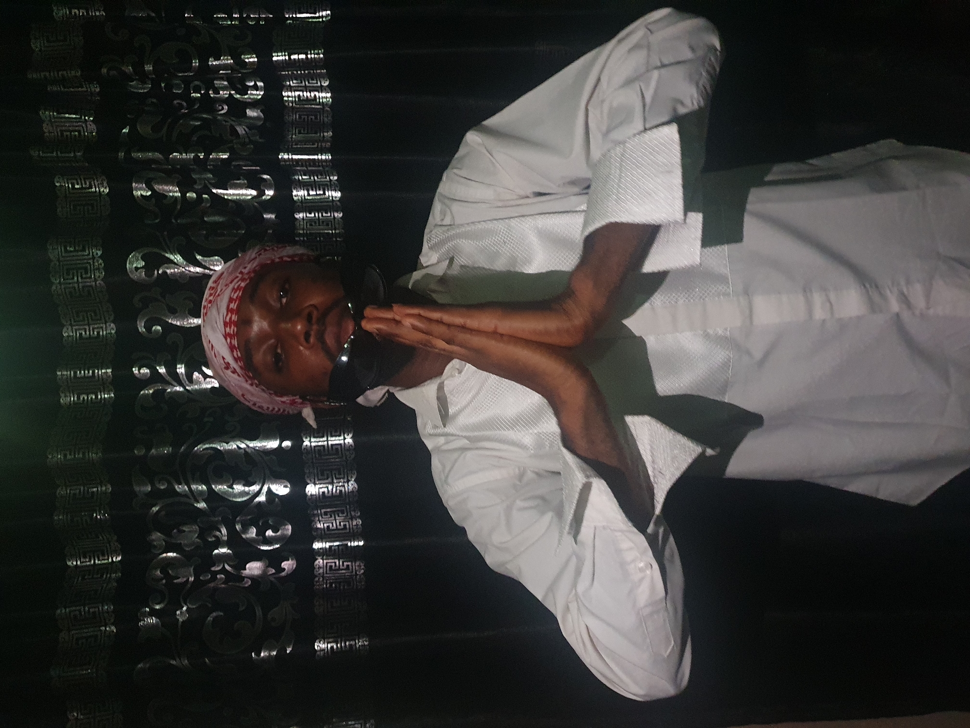A man wearing a white, button-down shirt and a red and white head covering is holding his hands together in front of his face. The man is standing in front of a black wall with ornate silver patterns. The image was taken indoors. It's impossible to ascertain if the image was taken in Lokoja, Nigeria, from the available information.