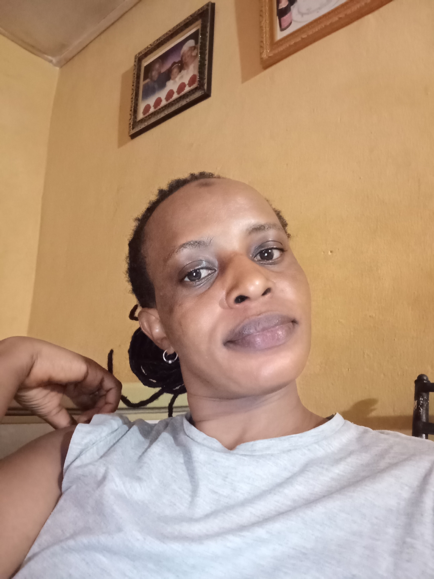A woman with dark brown skin and short dark hair braided behind her ear is shown in a close-up, angled selfie. She wears a light gray t-shirt and a small silver hoop earring. Her expression is neutral.
Behind her is a pale yellow wall with two framed pictures hanging high. The location is identified as Narayi, Nigeria.