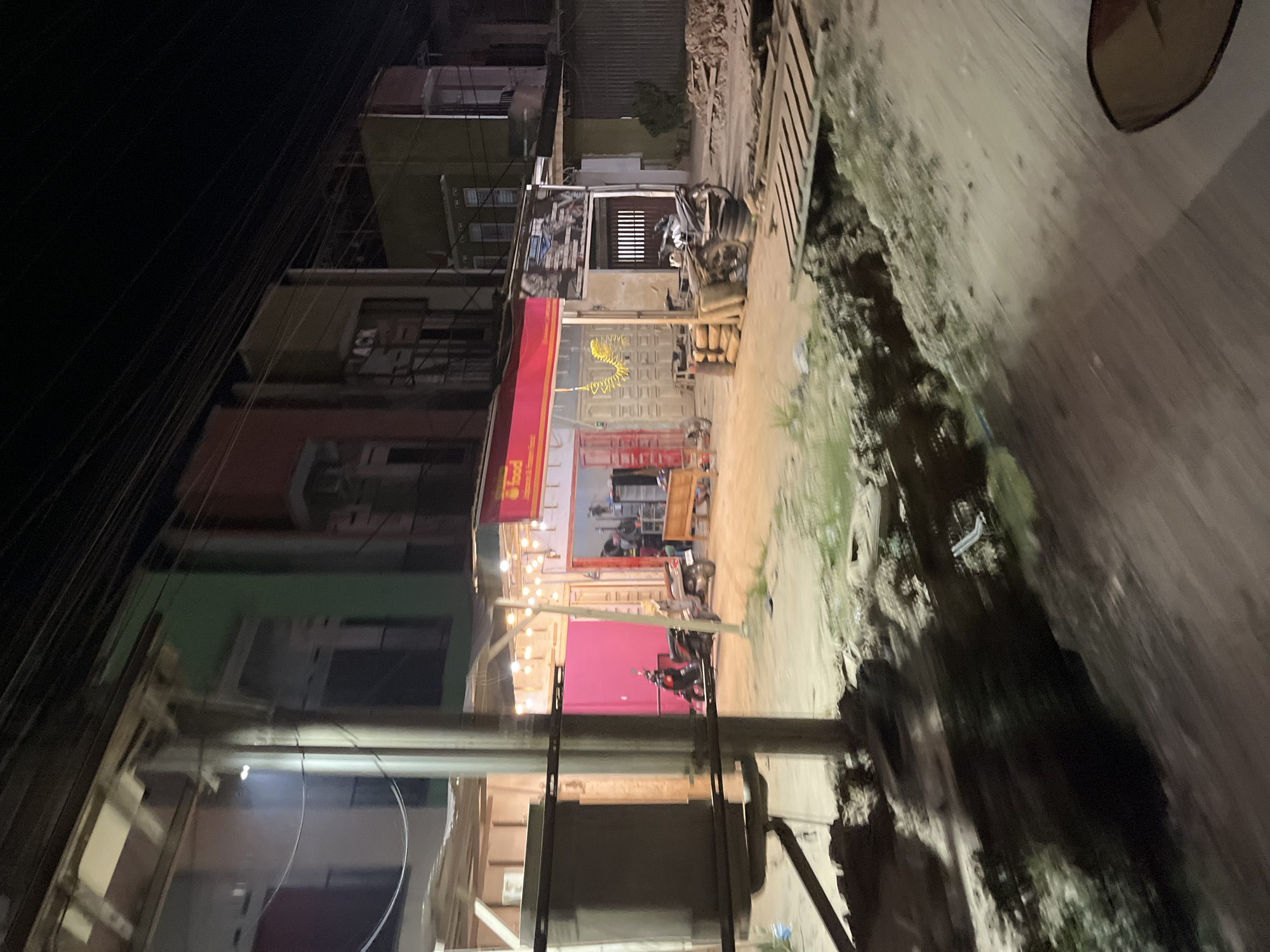 The image depicts a street scene at night in Benuaraja, Indonesia. The perspective is from a slight angle, making the buildings appear to be leaning. The buildings are multi-story structures, with some showing signs of construction or disrepair, indicated by exposed wooden beams and scaffolding. The ground level features shops or businesses. One shop has a red awning with some visible text that is difficult to read. Another establishment has a vibrant pink wall in the background, possibly a backdrop or a painted wall.
Illuminated by string lights, the street is partially visible. A motorcycle is parked in front of one of the shops, and another motorcycle is seen further down the street. Sandbags are stacked near one of the storefronts. The overall atmosphere is somewhat urban and developing, with a mix of commercial activity and ongoing construction. The time of day is clearly night, evidenced by the darkness and artificial lighting. The weather appears to be dry. There are no people prominently visible in the image. The scene is likely a commercial or mixed-use area within the Indonesian town.