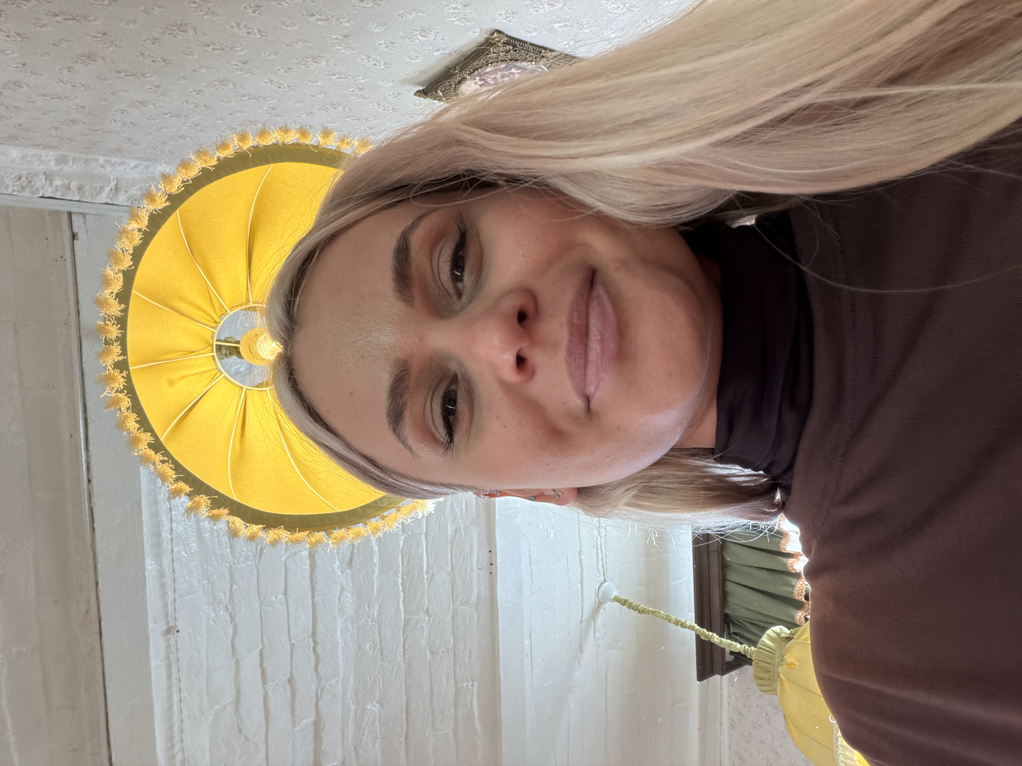 A selfie-style, upward-angle photograph features a blonde woman smiling slightly. She wears a dark brown turtleneck shirt. Behind her, a yellow lampshade with fringed trim is visible. The background includes a white brick wall and a patterned ceiling. There is a picture frame hanging on the ceiling. The location is not discernible as the city of Sarapul, Russia.