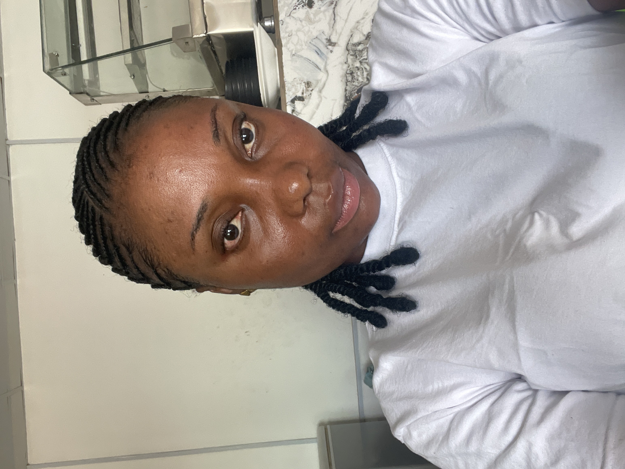 A close-up portrait shot, rotated 90 degrees clockwise, features a Black woman looking directly at the camera. Her face is well-lit, highlighting her dark skin, clear brown eyes, and a subtle, natural expression with a hint of a smile. Her dark hair is styled in cornrows across her scalp, with several short, dark twists or dreadlocks visible along the side of her head and near her shoulder. She is wearing a simple white t-shirt, which drapes loosely around her.
The background indicates an indoor setting in Jos, Nigeria. To the left of the woman's head (when oriented correctly), a plain, light-colored wall is visible. Above her and slightly to the right, a reflective metallic structure with glass elements, possibly a shelf or part of an appliance, can be seen. Further to the right and behind her, there's a surface with a mottled white and grey pattern, resembling a countertop or tiled wall. The lighting appears to be artificial and soft, illuminating the subject evenly. No text is visible in the image. The overall impression is a straightforward, casual portrait.