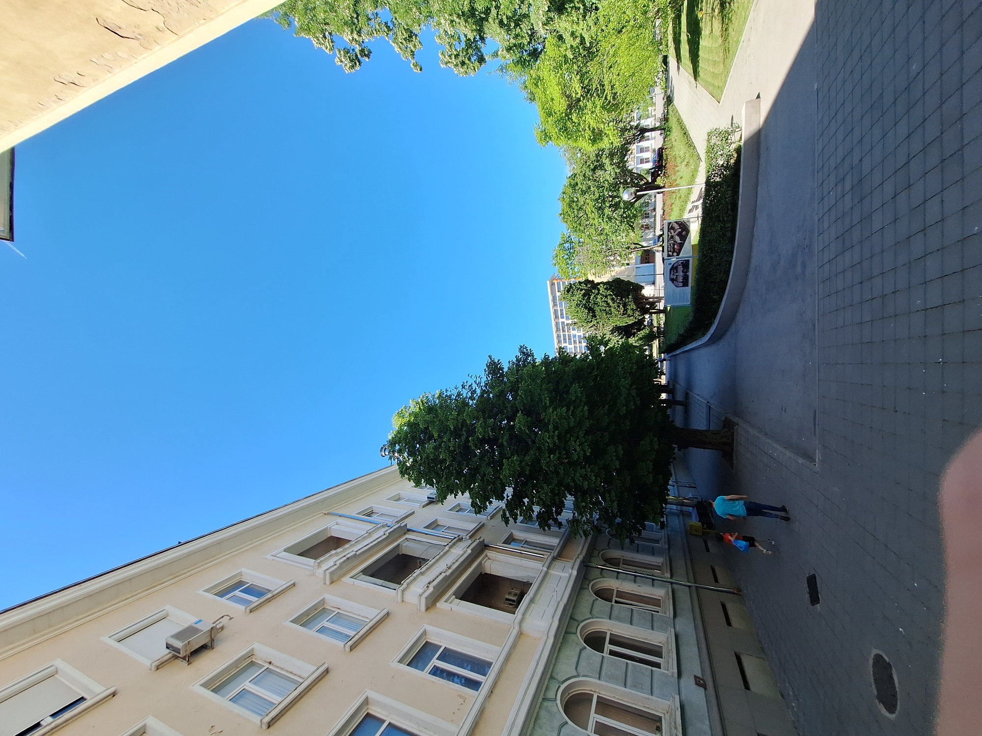 The image depicts an outdoor urban scene on a clear, sunny day, likely in the morning or early afternoon given the bright blue sky and strong shadows. The perspective is looking upwards and outwards, creating a slightly disorienting effect due to the image's rotation.
In the foreground, a light-colored building with multiple windows and balconies occupies the lower left portion of the frame. An air conditioning unit is visible on one of the walls. Adjacent to this building, on the right, is a paved sidewalk made of grey interlocking bricks.
Further into the scene, a lush green tree with a thick canopy stands prominently. Beyond the tree, a street or walkway curves, bordered by a strip of grass and more trees. Several other buildings, some appearing taller with many windows and others shorter, are visible in the background, interspersed with more greenery. A street lamp is present near the curved walkway.
Two figures are walking on the paved area. One person, wearing a blue t-shirt and dark pants, is walking away from the viewer. Another smaller figure, possibly a child, wearing an orange top and dark bottoms, is walking alongside. They appear to be casually strolling.
The overall atmosphere is one of a peaceful, everyday scene in a city park or residential area with pedestrian access. The clear weather and abundant foliage suggest a pleasant season, likely spring or summer. No visible text is discernible in the image.