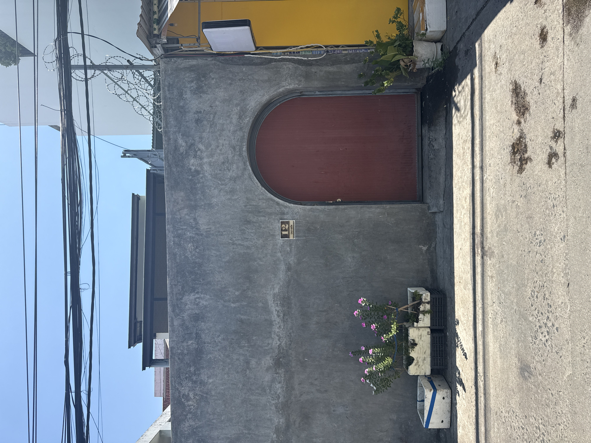 A sunny outdoor scene depicts a textured gray concrete wall with a dark red arched doorway. To the left of the doorway, a small potted plant with pink flowers is visible, alongside two white rectangular containers, one with a blue stripe, and two black crates. Above the plant, a small square sign with the number "12" and text below it is affixed to the wall. To the right of the doorway, a tall yellow building is partially visible, with a white square sign mounted on a pole next to the doorway. A large quantity of black electrical wires are strung across the top of the image against a clear blue sky, with barbed wire visible above the concrete wall on the right side. The ground in front of the wall is paved with concrete, showing some dark patches and shadows. The scene appears to be on a street in Bac Nha Trang Ward, Vietnam.