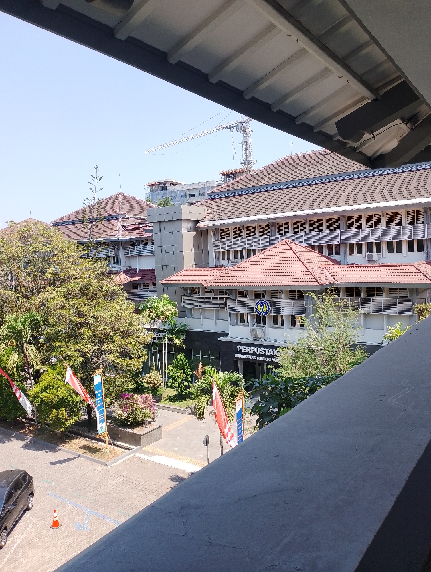 The image depicts the exterior of a building, likely a library or administrative building, in Depok, Indonesia. The building has a brown tiled roof and white facade with numerous windows. A prominent sign on the front of the building reads "PERPUSTAKAAN UNIVERSITAS NEGERI YOGYAKARTA". Several Indonesian flags are displayed in front of the building, along with trees and other vegetation. In the background, a construction crane is visible, suggesting ongoing development in the area. The scene is set against a clear blue sky. The perspective suggests the photo was taken from an elevated position, possibly from another building.