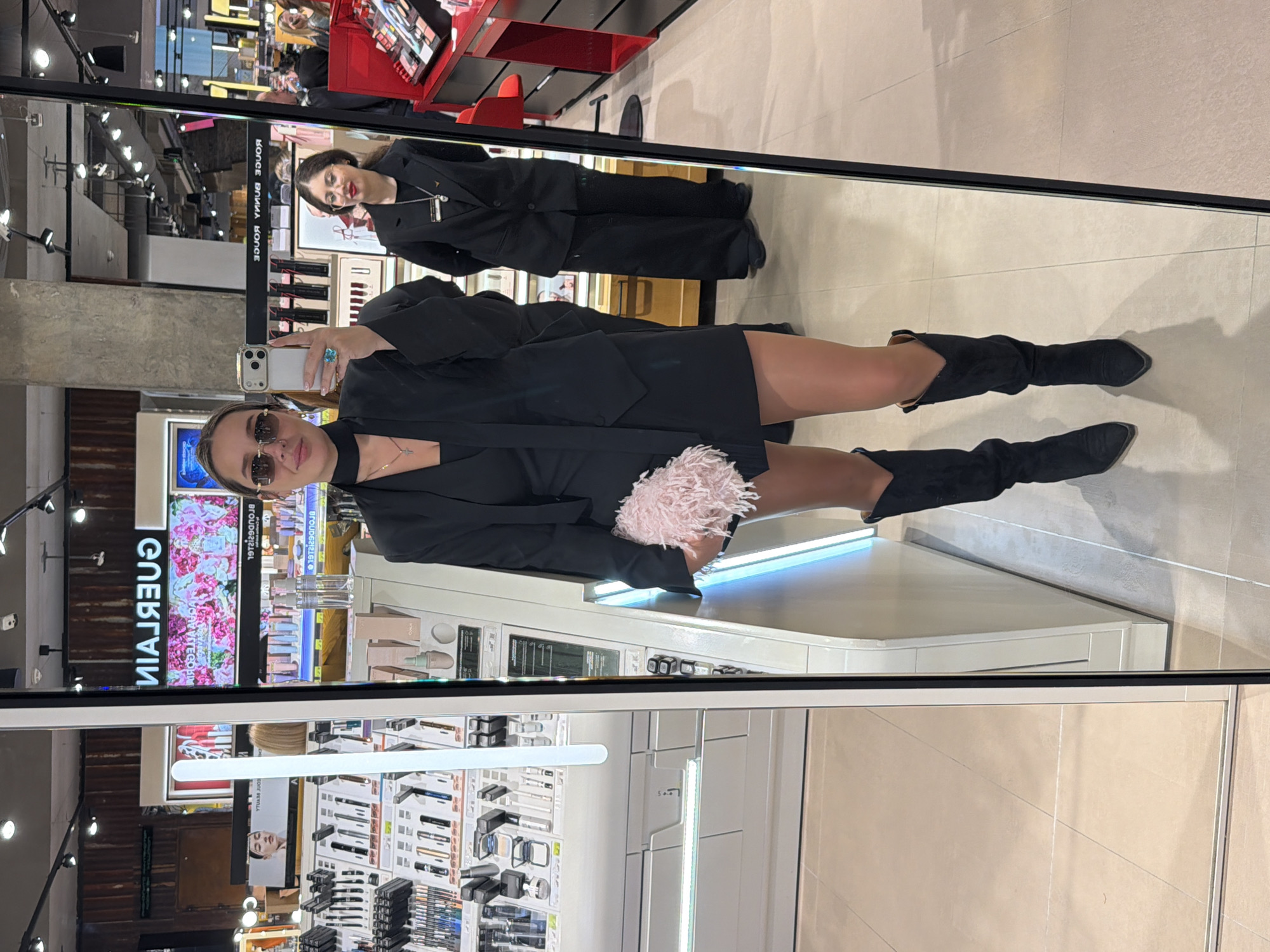 A mirror reflection shows a woman in a black blazer, a short black dress, and black knee-high boots holding a phone and standing in what appears to be a beauty or department store. The reflection shows rows of cosmetics and other beauty products on display behind her. A second person is seen in the reflection wearing a similar outfit. The setting is indoors, illuminated by artificial lighting. The location is potentially Moscow, Russia, but there's no definitive evidence within the image to confirm this.
