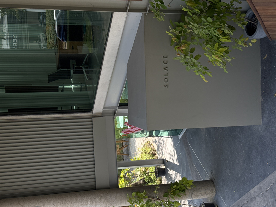 An exterior view shows a modern building with a large window and a sign reading "SOLACE." The building features metal siding, and a concrete awning hangs above the entrance. Lush green plants are present on the side. A view of a patio with furniture is visible through the awning. The ground is paved, and the image appears to be taken from an angle.