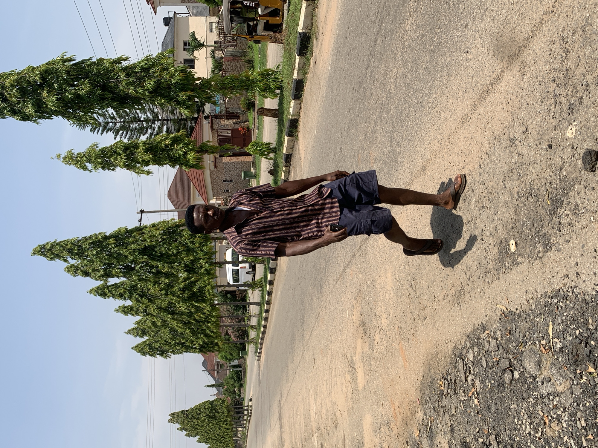 A young Black man walks on a paved road in Abuja, Nigeria. He is wearing a striped short-sleeved button-down shirt and dark blue shorts. He is also wearing flip-flops and carrying a mobile phone in his right hand. The road is lined with tall, slender trees on both sides. In the background, there are houses, a white van, and a yellow auto-rickshaw. The sky is clear and blue. The road is in a residential area with well-maintained properties.