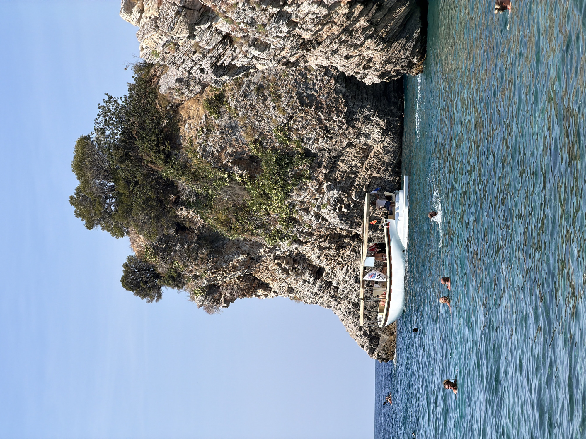 A boat tour is underway near a rocky cliff face in Budva, Montenegro. The boat, a white vessel with a covered seating area, is anchored near the cliff. Several people are visible on the boat, some seated and some standing. The water is a clear turquoise and several individuals are swimming and enjoying the sea. In the distance, a person in a kayak is paddling. The rocky cliff is covered in sparse vegetation and features distinct layers. The sky is clear and blue.