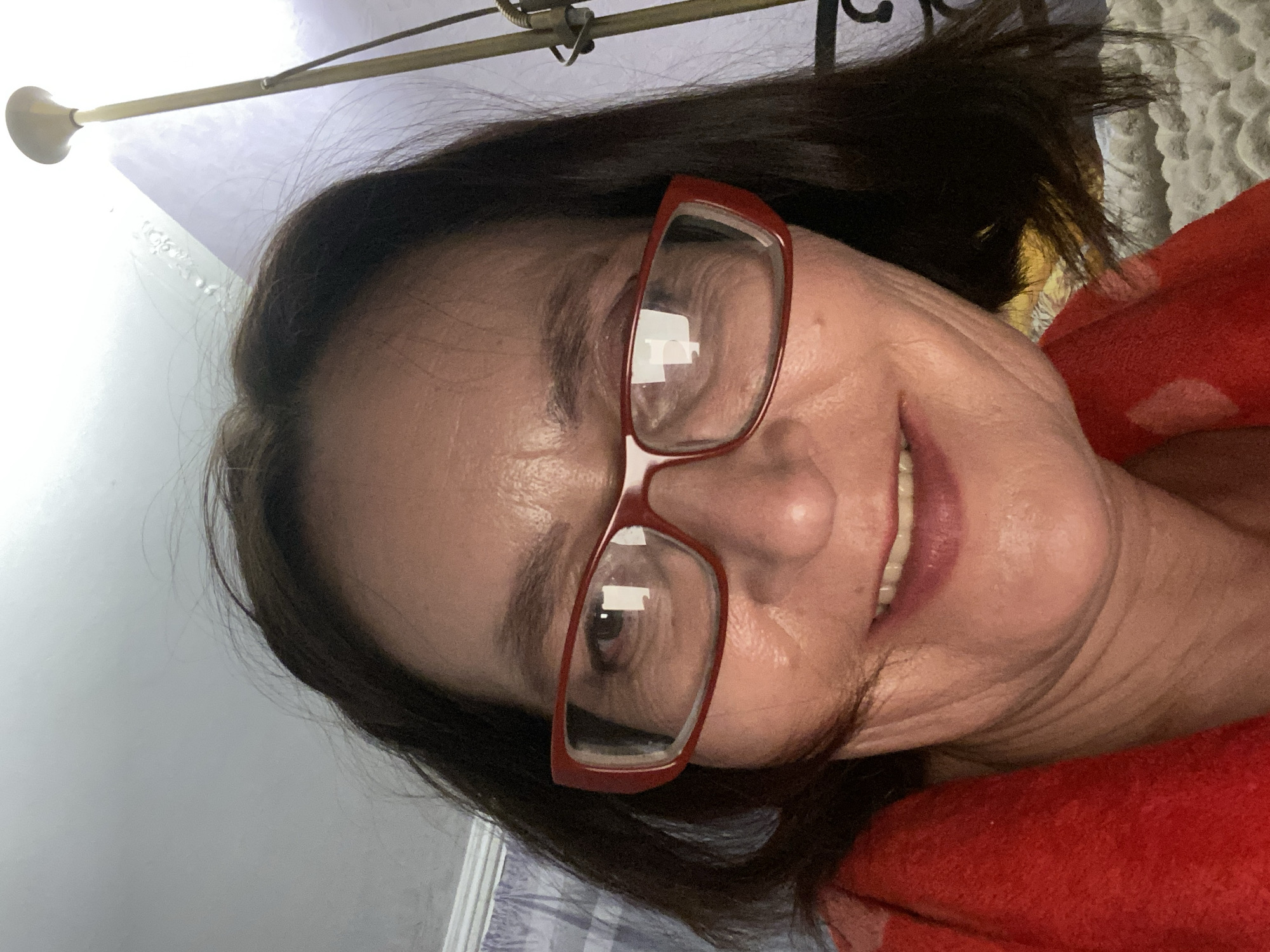 A woman with dark hair and red glasses is smiling at the camera. She is wearing a red garment. The background includes a bed with patterned linens and a light-colored wall. A lamp is visible in the upper left corner. There is no information to indicate this image was taken in Novokuznetsk, Russia.