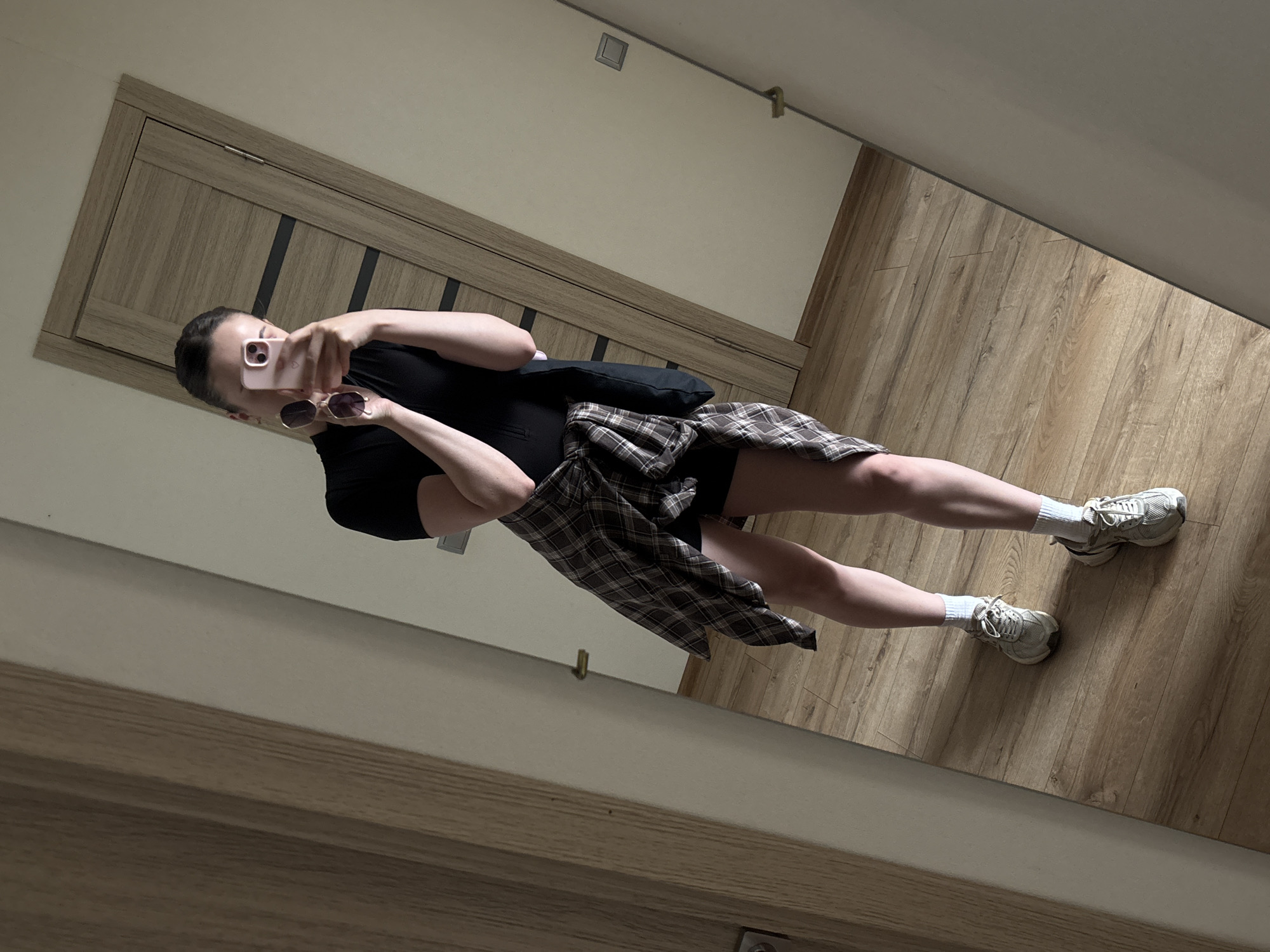A woman takes a mirror selfie indoors. She is wearing a black t-shirt dress, a plaid shirt tied around her waist, white socks, and athletic shoes. She is holding sunglasses and a pink smartphone in her right hand. The room has wooden flooring and a wooden door in the background.