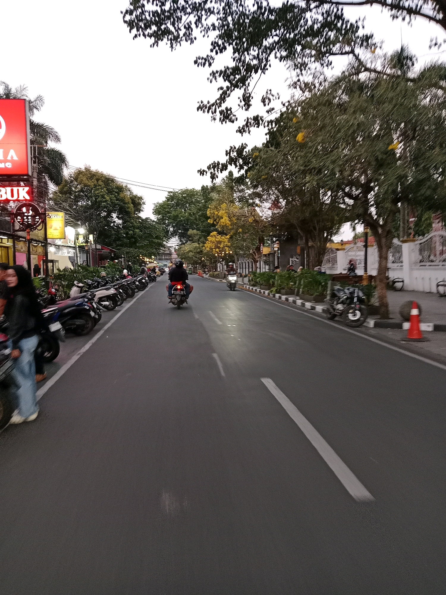 The image shows a street scene in Cianjur, Indonesia, at dusk. A paved road runs diagonally from the lower right to the upper left of the frame. On the left side of the road, a row of parked motorcycles is visible, with a woman in a black jacket and blue jeans standing next to them. In the background on the left, there are buildings with neon signs, including a red sign with white text and a black sign with illuminated text. Trees line the right side of the road, some with yellow flowers. Several people are on motorcycles, one of whom is riding away from the camera down the street. An orange traffic cone is placed on the sidewalk on the right side of the road. The sky is overcast.