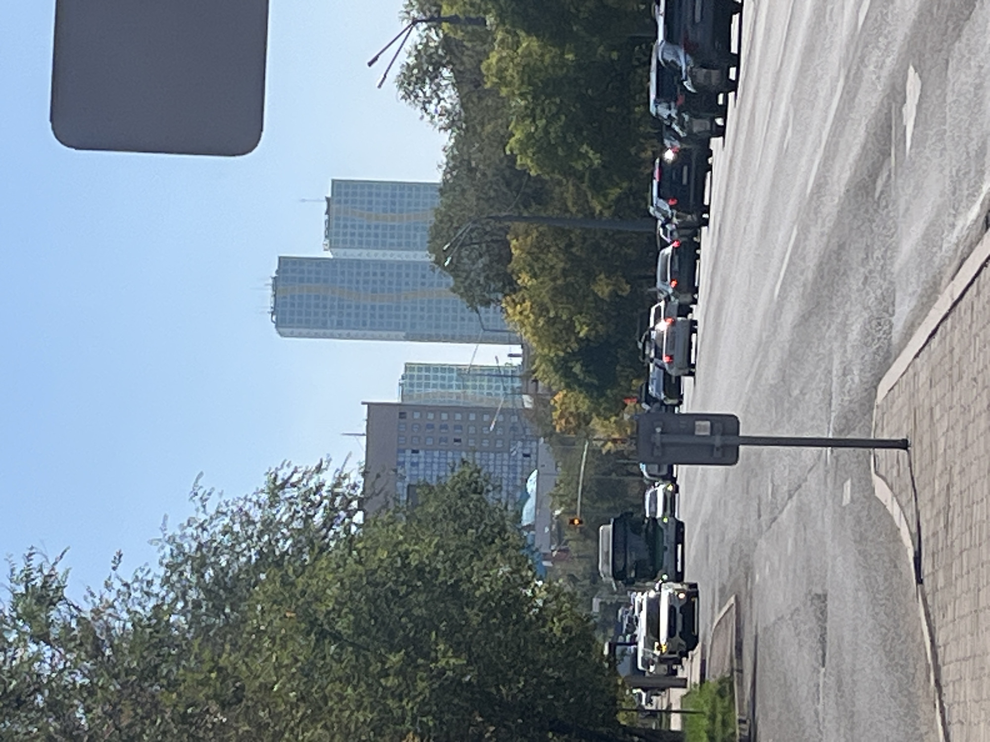 The image depicts a wide, paved road in Astana, Kazakhstan, on a clear, sunny day. Tall, modern buildings with glass facades dominate the background, including at least two prominent skyscrapers. Trees line the sides of the road, with some foliage showing autumn colors. Traffic is moderate, with several cars and a bus visible on the road. The perspective is from street level, likely from a sidewalk, showing a pedestrian area with brick paving in the foreground. A traffic light is visible, showing red. A grey signpost is positioned on the right side of the road. The scene appears to be a typical urban environment in Astana, characterized by a mix of greenery and contemporary architecture.
