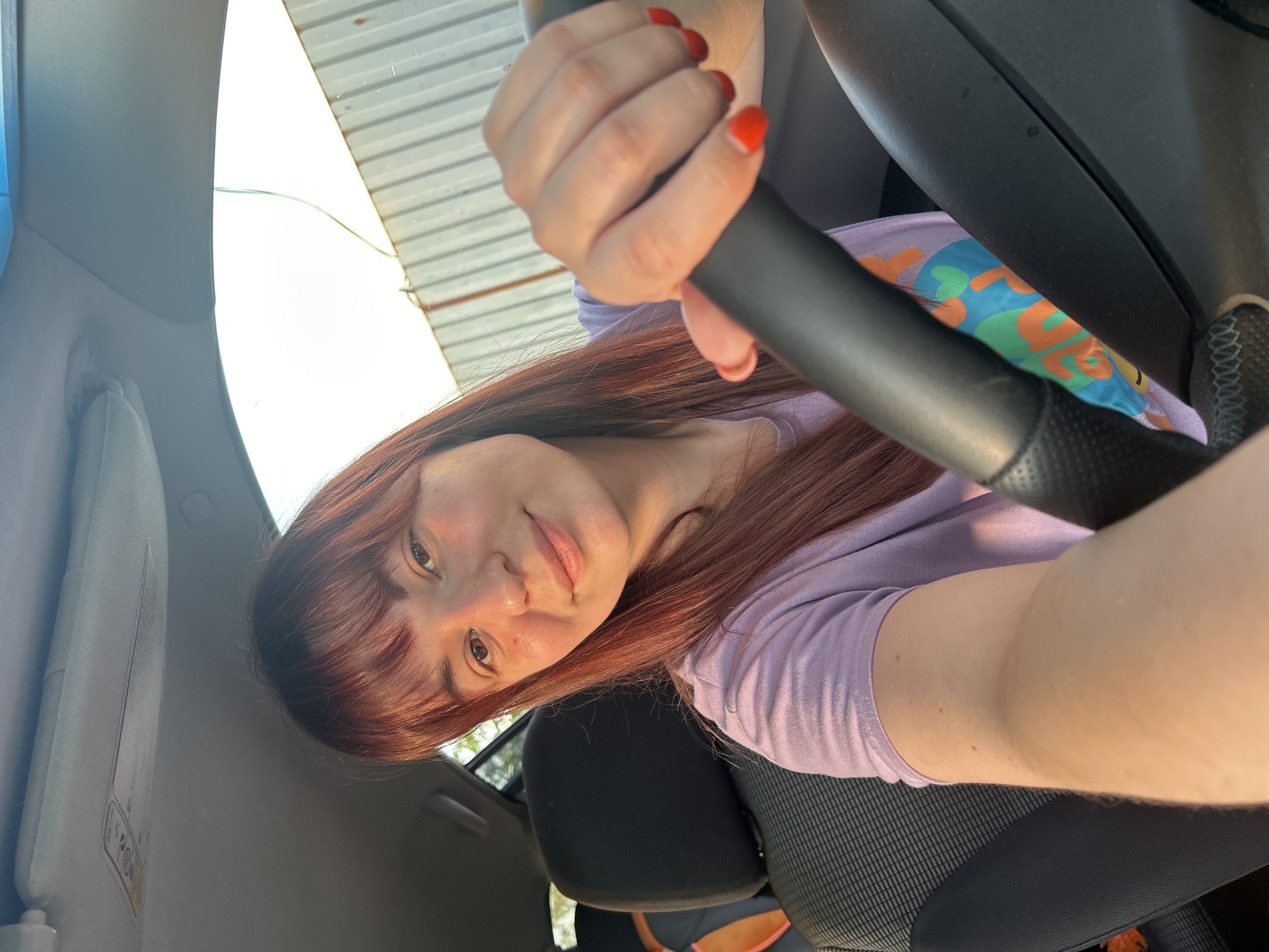 A woman with reddish-brown hair and red nail polish is holding a black steering wheel inside a car. She is wearing a light purple t-shirt with a colorful print. The interior of the car includes a black seat and a visor on the left side. Through the windshield, a glimpse of a metal structure can be seen. The image provides insufficient evidence to determine the location in the city of Фоки, Russia.