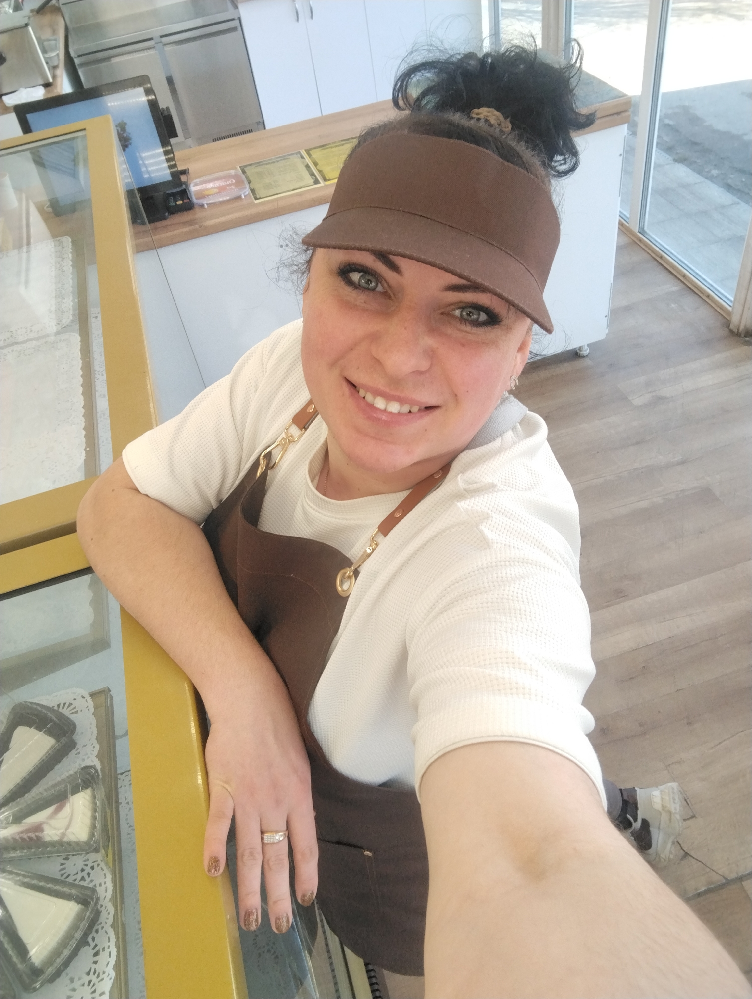 A medium-shot, high-angle selfie photograph depicts a smiling woman indoors. She is wearing a brown visor, a white waffle-textured top, and a brown apron with tan leather straps and brass hardware. Her hair is pulled up into a messy bun. She is resting her hand on a glass display case with a gold-colored frame that contains several pre-packaged triangular slices of cheesecake. A wedding ring is visible on her left ring finger.
In the background, there is a cash register system on a counter and white cabinets and stainless steel appliances. A window is visible behind her, revealing an exterior scene.
The photograph was taken in Votkinsk, Russia.
