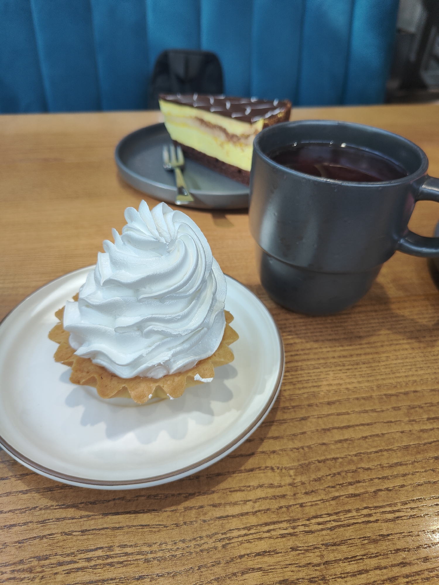 An overhead medium shot captures a dessert and drink set on a wooden table. In the foreground, a white meringue tart sits on a white plate with a brown rim. Behind it, a slice of chocolate and yellow layered cake rests on a dark grey plate, with a silver fork on the plate. To the right, a dark grey mug filled with dark liquid, likely tea or coffee, is positioned on the table. The background features a blurred teal cushioned bench and a hint of a black chair. The scene is set in a cafe, likely in the city of Chornomorsk, Ukraine.