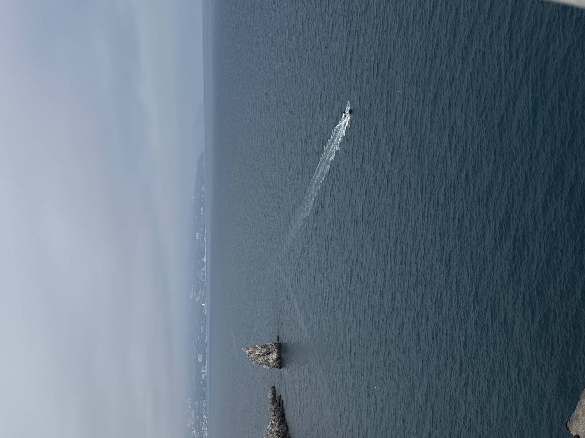 The image shows an aerial view of the Black Sea off the coast of the Гаспринский поселковый совет, Ukraine. In the foreground, there are two rocky outcroppings protruding from the water. A boat is moving through the sea, leaving a white trail of wake. In the background, the hazy coastline of the town can be seen.