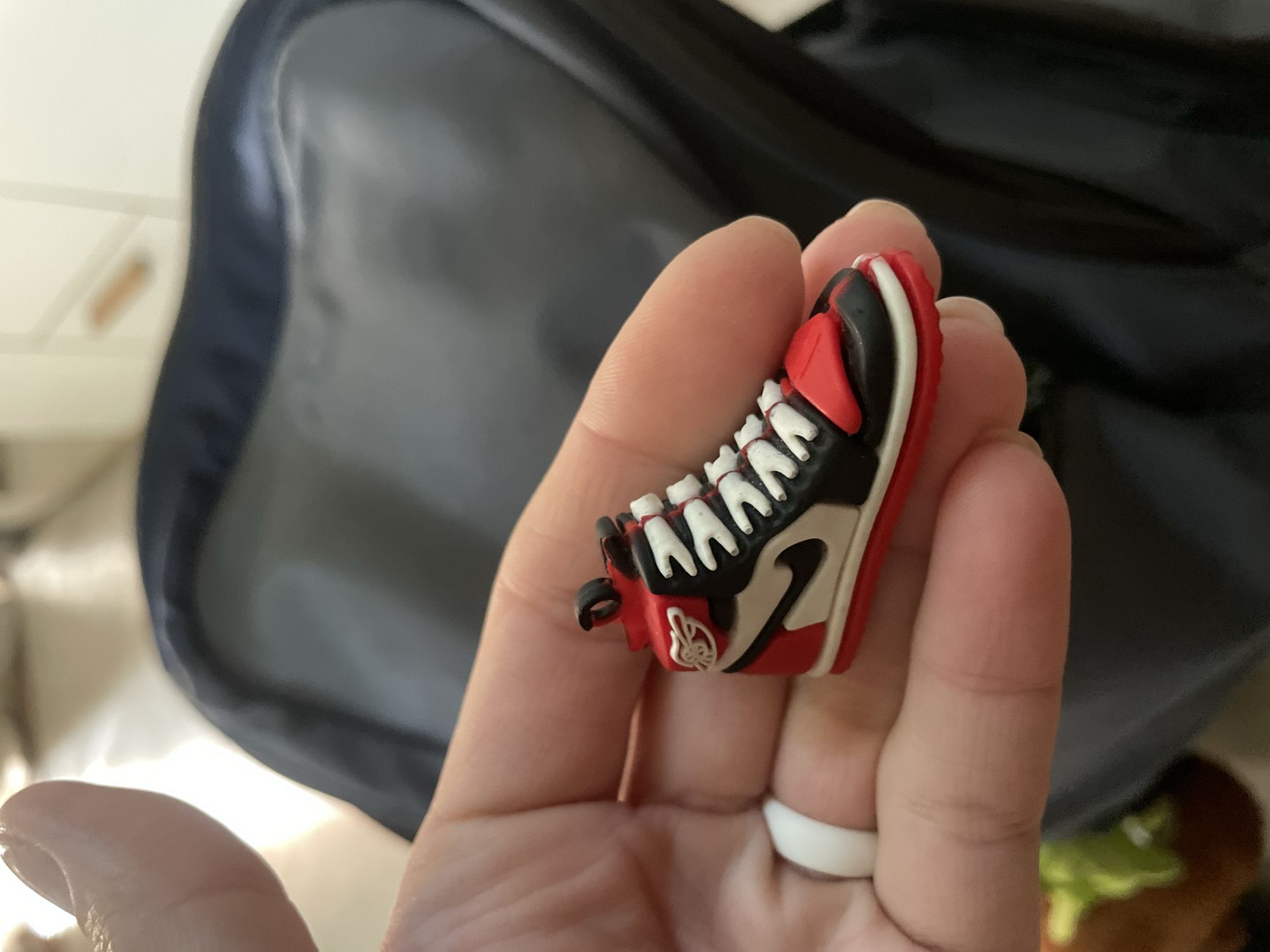 A close-up shot features a human hand holding a miniature sneaker. The hand has light-toned skin and a white ring is worn on the ring finger. The small sneaker is highly detailed, resembling an Air Jordan 1, predominantly colored in red, black, and white, with visible white laces and a small black Nike swoosh on the side. A white "Wings" logo is also discernible on the ankle area of the miniature shoe. The background is out of focus, showing a large, dark-colored object, possibly a bag or piece of luggage, behind the hand. To the left, a lighter, indistinct surface can be seen. In the lower right corner, a blurry object with green and brown tones is vaguely visible. The image captures the act of presenting or examining this small, collectible item in an indoor setting. No specific time of day, weather, or emotions are discernible, and the location context of Baranovka, Russia, is not visually identifiable within the image.