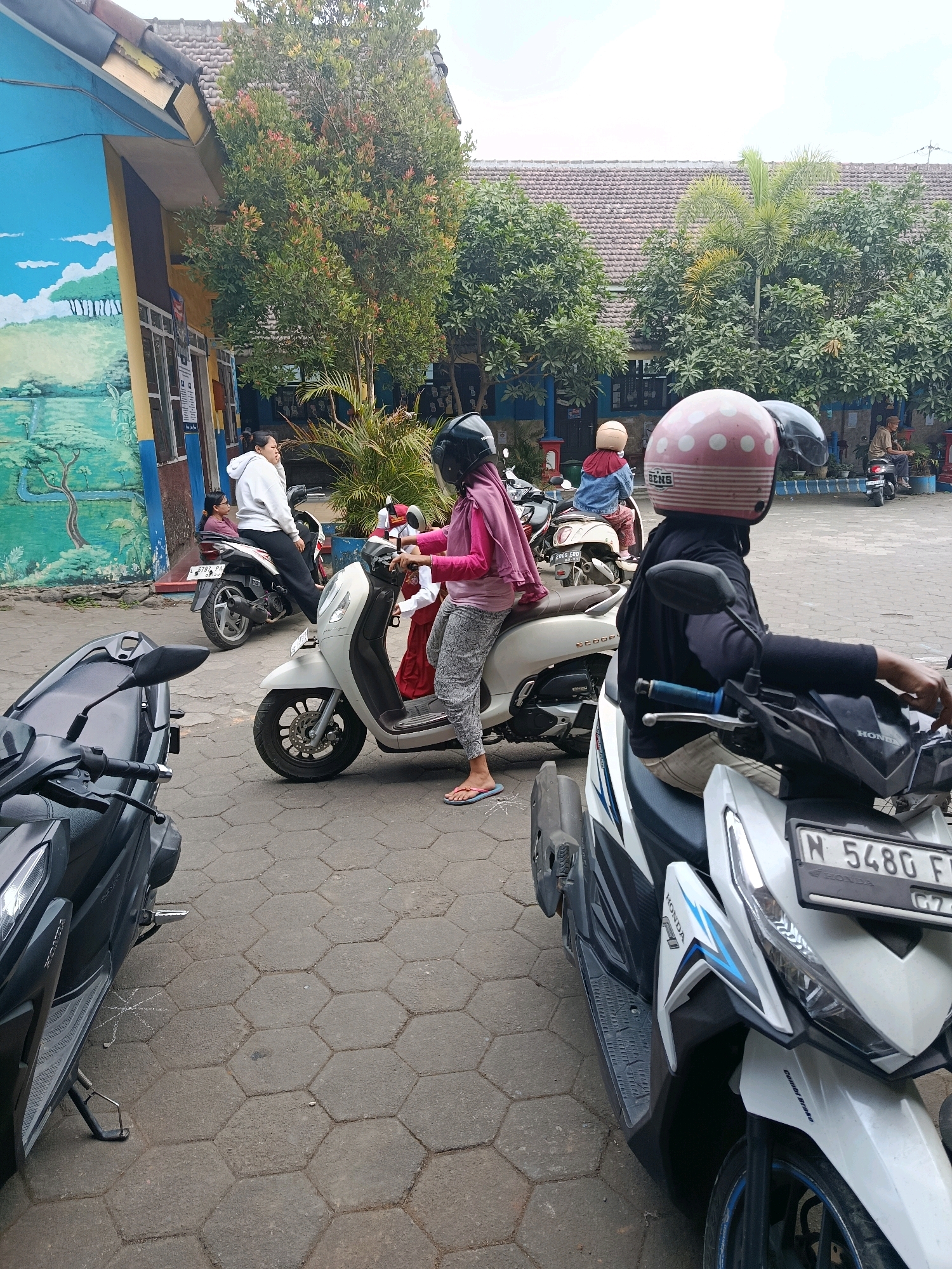 In Bululawang, Indonesia, a group of people are gathered around several parked scooters outside a building with a mural on its wall. A woman wearing a pink hoodie and grey patterned pants, with a pink helmet adorned with white polka dots, sits on a white scooter. She appears to be holding onto the handlebars, looking towards the left. Another woman, wearing a white hooded sweatshirt, is seated on a white scooter in the background, facing away from the camera and looking to her left. A third person, wearing a blue jacket and a beige helmet, is also on a scooter behind the woman in the white sweatshirt. In the foreground, a black scooter is parked, facing left, with its front wheel and part of the body visible. The ground is paved with hexagonal concrete tiles. Trees with green foliage are visible behind the building and scooters, and the sky is overcast. The building has a tiled roof and a blue and yellow exterior wall with a mural depicting a landscape.