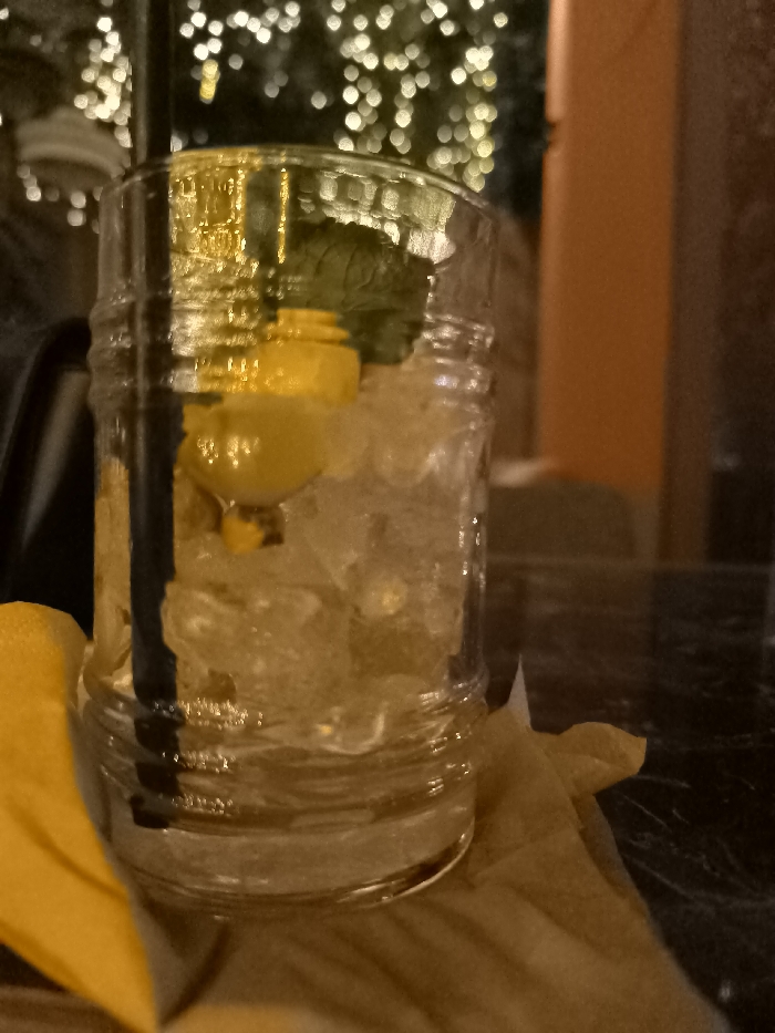 This close-up image features a refreshing drink served in a clear, textured glass, possibly a mason jar style. The glass is filled with crushed ice, a prominent black straw, a bright yellow lemon wedge or slice, and a sprig of green mint, suggesting a cocktail or iced beverage. The glass rests on a yellow paper napkin or coaster, which is placed on a dark, reflective table surface, possibly marble or polished wood.
The setting is an outdoor or semi-outdoor area at night, likely a bar, restaurant, or cafe patio in Palaio Faliro, Greece. The background is intentionally blurred, creating a bokeh effect from numerous warm, circular lights, indicative of decorative string lights on trees or surrounding structures, contributing to a festive and relaxed ambiance. A dark vertical element, possibly a pole or support, is visible to the left of the glass, while a blurred, orange-brown vertical surface, perhaps a building wall or furniture, is present on the right side of the frame.
The image captures a moment of leisure, emphasizing the drink itself. The time of day is clearly nighttime due to the artificial lighting and overall dark environment. No people are visible, and there is no discernible text within the image. The scene conveys a calm, inviting atmosphere.