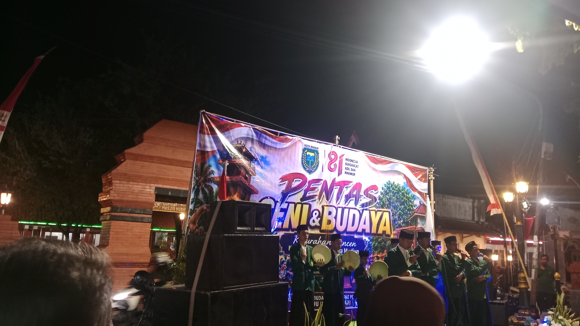 The image depicts an outdoor event at night, likely a cultural performance, in front of a brick structure that appears to be the entrance to a mosque, indicated by Arabic script and "MASJID NURHI" on a sign. A large banner with the text "PENTAS SENI & BUDAYA" (Art & Culture Performance) and "KOTA MADIUN" (Madiun City) is prominently displayed. The banner also features the number "81" and the slogan "INDONESIA BERDAULAT ADIL DAN MAKMUR" (Indonesia Sovereign Just and Prosperous).
Several individuals dressed in green robes and black caps are on a stage, some holding circular percussion instruments and microphones, suggesting they are performers. A stack of large speakers is positioned to the left of the stage. The background shows trees, streetlights, and architectural elements, all illuminated by artificial lighting. The overall atmosphere suggests a local celebration or festival in Madiun, Indonesia.