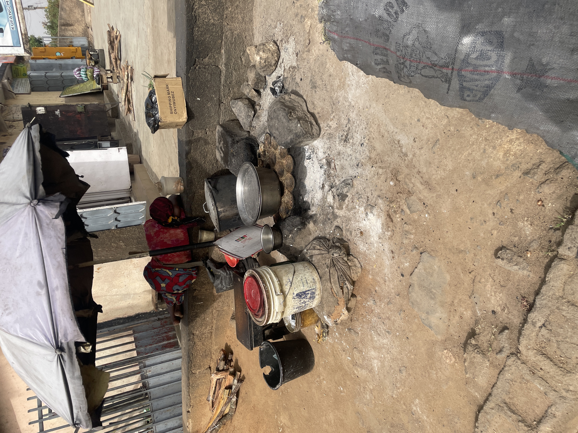 This image depicts an outdoor cooking scene in Jos, Nigeria. A woman in a red and blue patterned garment sits next to a makeshift cooking area. Several cooking pots sit atop a fire pit constructed from rocks. A black bucket and a paint bucket rest beside the fire. Firewood is scattered nearby. An umbrella provides shade. The ground is a mix of dirt and concrete. A black tarp or similar material partially covers the upper right corner of the scene. In the background, there are stacks of metal materials and a box with printed text.