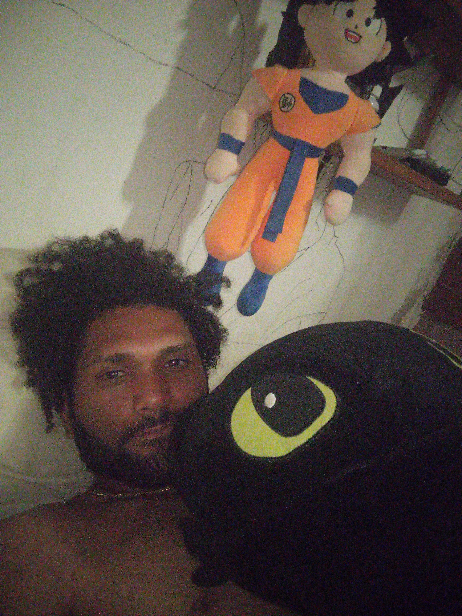 A man with dark curly hair and a beard lies on a surface, holding a black plush toy with a yellow eye. Behind him on the same surface is a plush toy resembling the character Goku from Dragon Ball, wearing an orange gi. The background appears to be a wall. The image provides no information to indicate that the location is El Valle del Espíritu Santo, Venezuela.