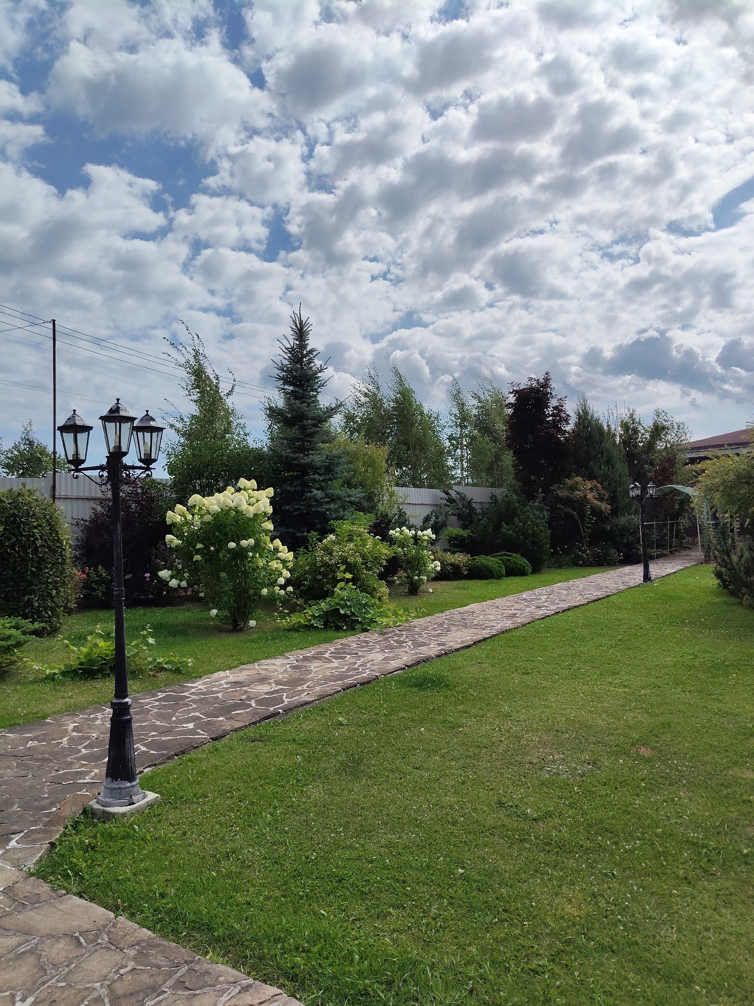 The image depicts a paved garden path winding through a lush green lawn. The path, made of irregular stone slabs, curves to the right, leading into a landscape dotted with various trees and shrubs. In the foreground, a black, three-headed lamppost stands to the left of the path. Further along the path, another similar lamppost is visible. The garden is characterized by a variety of greenery, including a large white flowering bush, evergreen trees, and leafy bushes. The sky above is a mix of blue and white clouds. The setting appears to be a private garden or park. The image provides no specific information about the city of Bogdanovka, Russia.