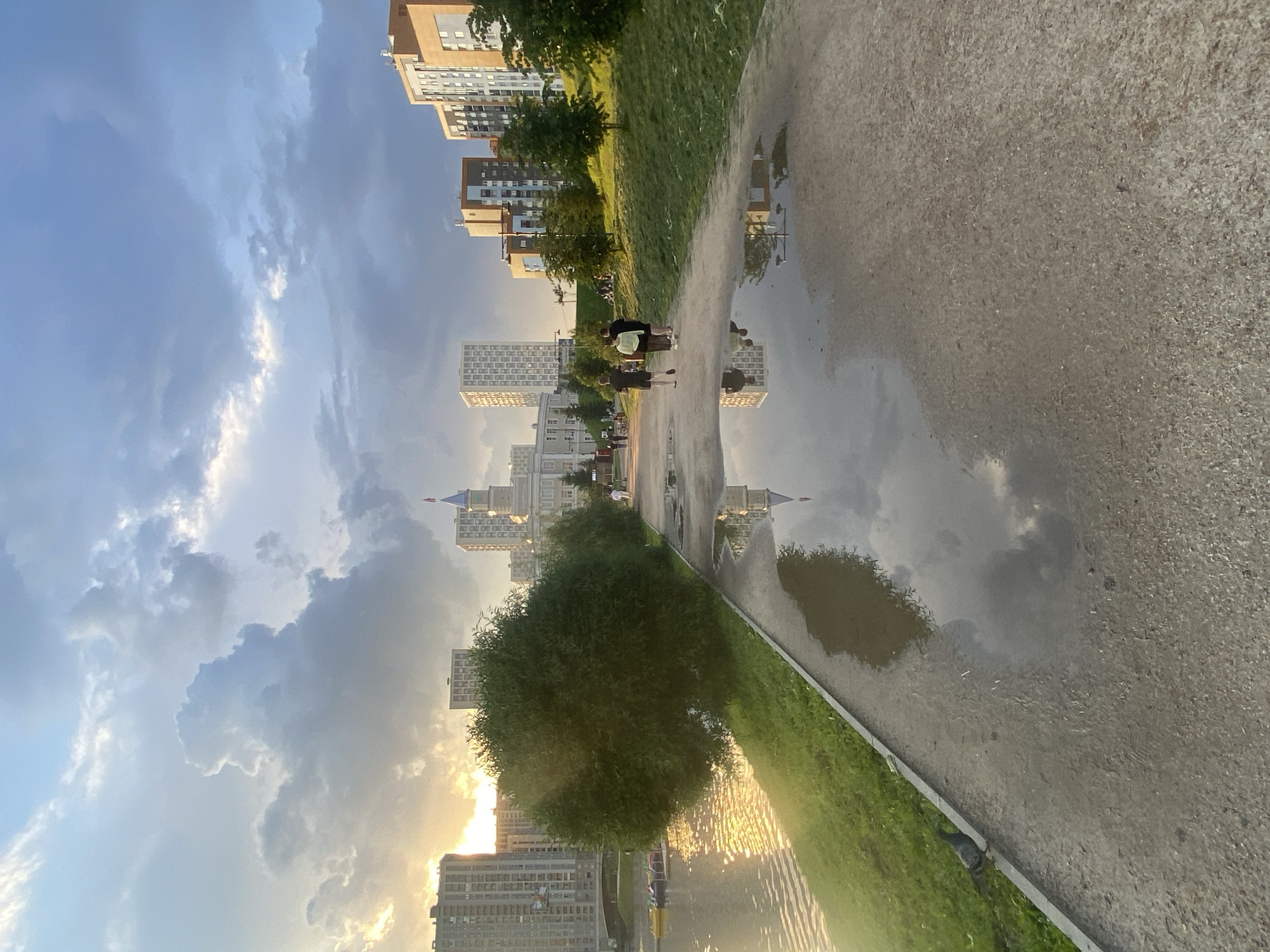 The image depicts a waterfront promenade in Yekaterinburg, Russia, likely the embankment of the Iset River, as evidenced by the surrounding urban landscape featuring apartment buildings and a prominent domed structure. The time of day appears to be late afternoon or early evening, with the sun casting a warm glow and creating reflections on the water. The sky is dramatic with large, cumulus clouds, some illuminated by the setting sun.
In the foreground, a paved walkway runs alongside the river. Several puddles are visible on the path, reflecting the sky and surrounding buildings. A lush green grassy area lines the left side of the walkway, bordering the water. On the water, several small, colorful boats are moored.
Further along the walkway, three individuals are walking away from the camera. Two are in dark clothing, and one is wearing a lighter colored top. Behind them, more people can be seen in the distance, engaged in leisure activities.
The background is dominated by a series of multi-story apartment buildings, varying in architectural style and color. A distinctive building with a domed roof and spire is visible, possibly a government building or a historical landmark. Trees are interspersed throughout the scene, adding to the park-like atmosphere. A pigeon is visible on the edge of the walkway, near the grass. The overall scene suggests a well-maintained urban park area enjoyed by residents for recreation.