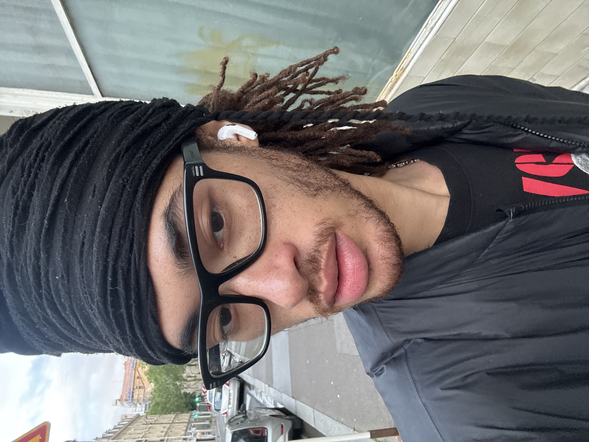 A close-up, tilted portrait of a man in an urban setting. He is wearing a black head wrap and rectangular black-framed glasses. He has dreadlocks and is wearing an earbud in his left ear. He is wearing a black jacket over a black t-shirt with some red writing. The background shows a street scene in Paris, France with buildings and cars.