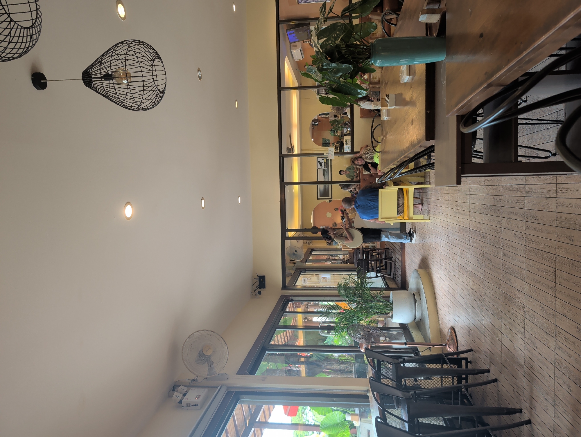The image depicts an indoor scene in Thong Sala, Thailand. A man in a blue shirt is playing a musical instrument in a cafe-like setting. Behind him, a woman is sitting and observing. The room features wooden floors and a long wooden table with chairs. Plants are present both indoors and visible through the windows, which offer an outdoor view. Lighting fixtures are mounted on the ceiling, and a fan is affixed to the wall near the window.