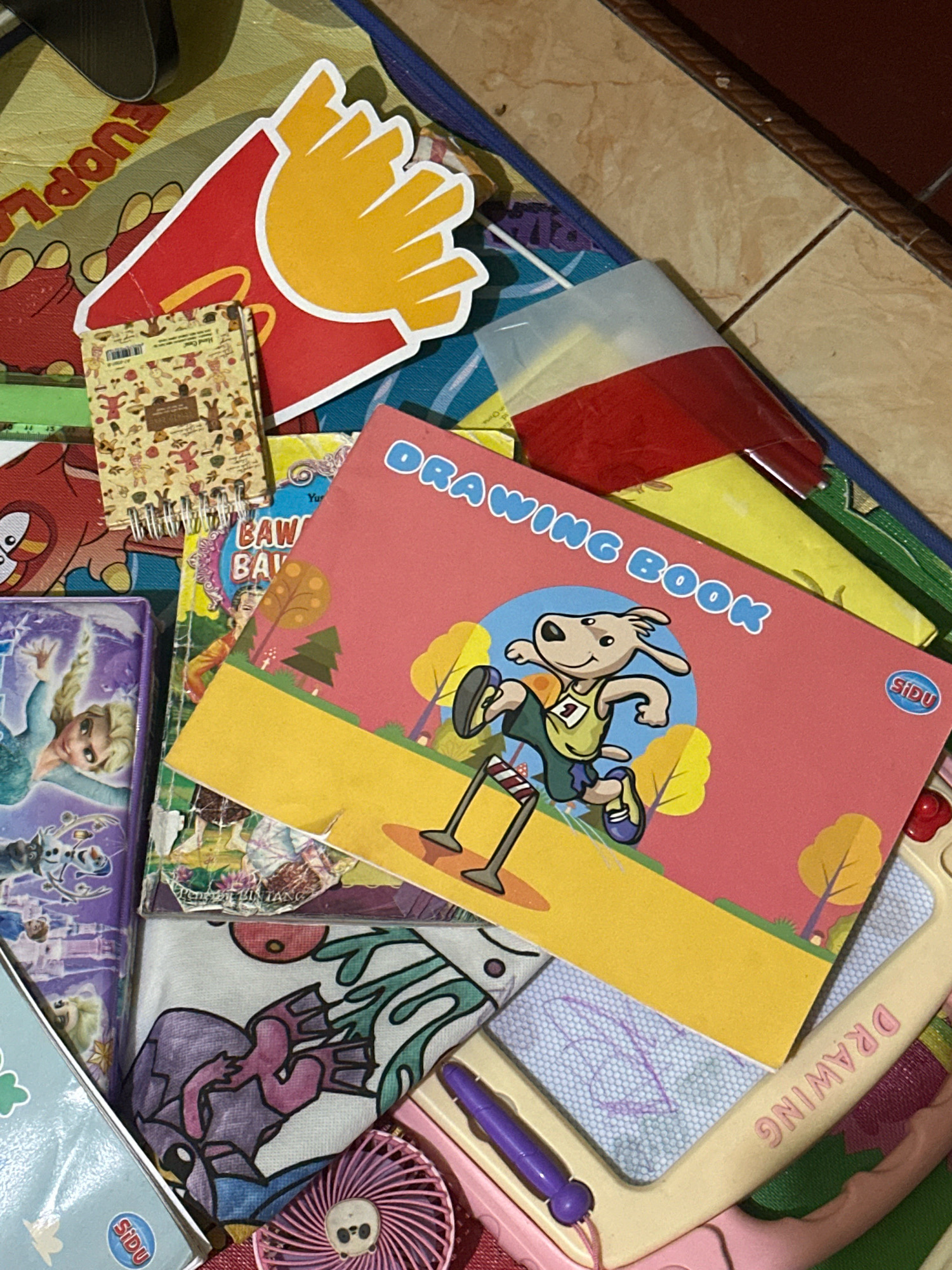 The image shows a collection of items on a surface, likely a floor, with a tiled background visible in the upper right corner, suggesting an indoor setting. The items include several books and what appears to be a drawing or writing pad. One prominent item is a pink book with the words "DRAWING BOOK" in blue lettering. This book features a cartoon dog character participating in a race, with a stylized landscape in the background. Next to this, a smaller notebook with a patterned cover is visible, along with a red and yellow cardboard cutout resembling french fries. To the left, a purple-covered item features a character that resembles Elsa from Disney's Frozen. Other items include a white cloth with a colorful design, a pink fan, and a drawing board with writing on it. The overall impression is of a child's activity area or a collection of art supplies. The environment does not provide specific clues to determine the exact location within Ngasem, Indonesia.