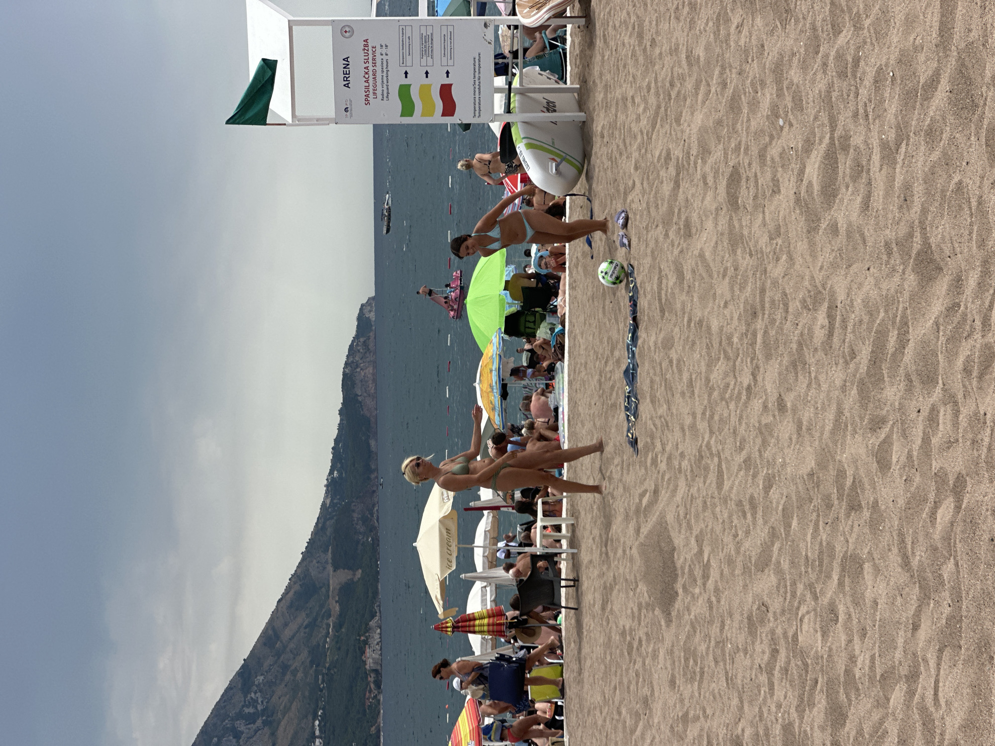 A group of people are enjoying a sunny day at the beach in Boreti, Montenegro. The scene is set on a sandy beach with the sea and a mountainous coastline in the background. The weather appears to be warm and clear, with a bright sky.
Several beach umbrellas and chairs are scattered across the sand, providing shade for the beachgoers. In the foreground, two women in bikinis are captured in mid-action, seemingly playing or posing. One woman, with blonde hair, stands gracefully, while another woman with dark hair is in a dynamic pose, her body in motion. A small, green soccer ball is visible on the sand near them.
Behind them, more people are relaxing and enjoying the water. Umbrellas of various colors, including a bright green one, dot the scene. A surfboard is propped up near a lifeguard station. A sign with text in Croatian and English is prominently displayed, indicating "ARENA SPASILAČKA SLUŽBA LIFEGUARD SERVICE" and providing working hours and information about sea and air temperature. The text also mentions "ice cream," suggesting a vendor is nearby. The overall atmosphere is one of leisure and summer enjoyment.