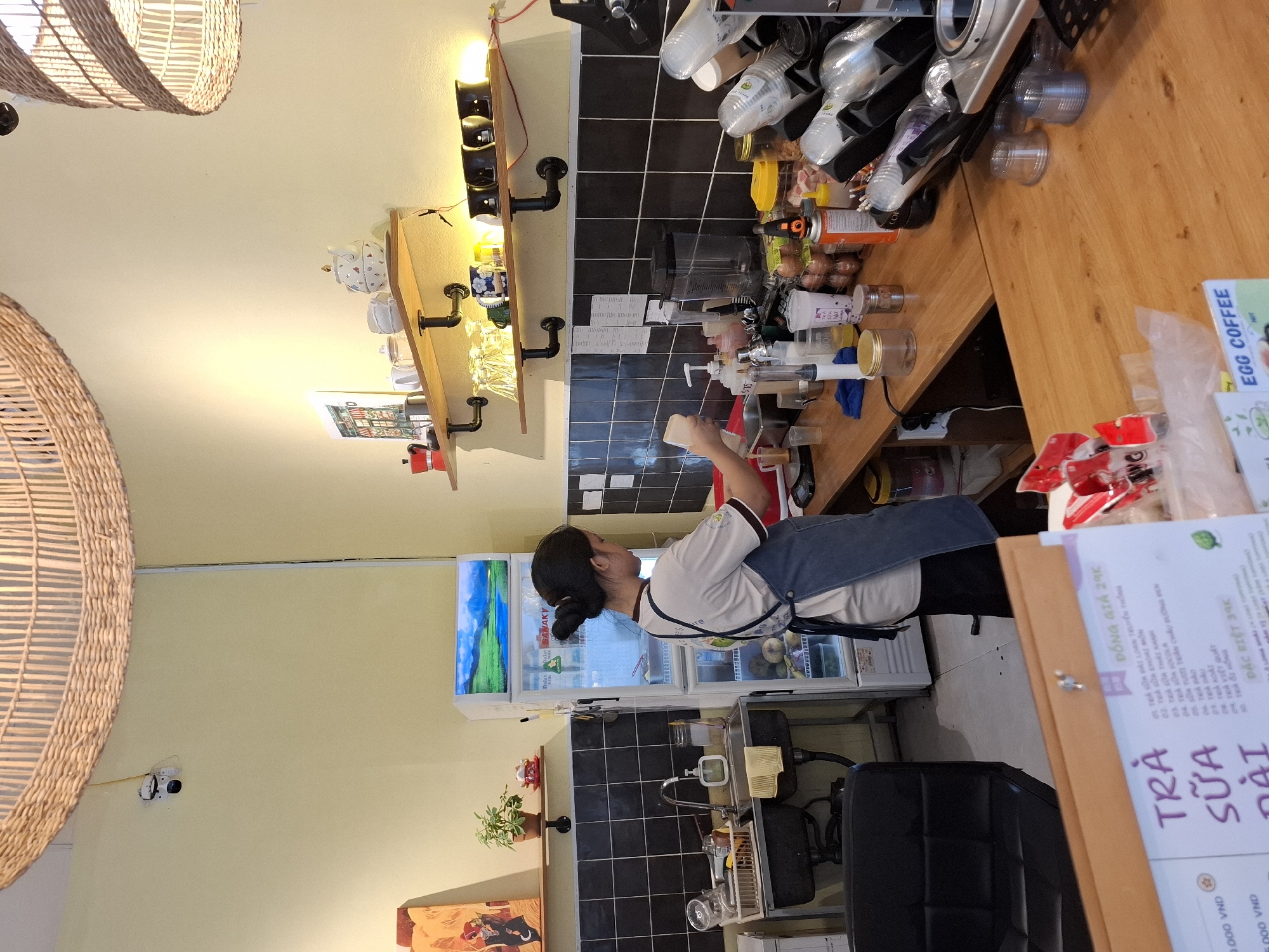 The image depicts the interior of a cafe in Nha Trang, Vietnam. A young woman with her hair in a bun, wearing a white t-shirt and a dark blue apron, is standing behind a wooden counter. She is in the process of preparing a drink, holding a clear plastic cup and a bottle of liquid.
The counter is cluttered with various items, including bottles of syrups, cups, a blender, and containers of ingredients. To the left of the counter, there is a stainless steel sink with a faucet and a dish rack filled with glasses. A white refrigerator with a landscape image on its door is situated next to the sink. Above the counter, shelves are mounted on the wall, displaying decorative teapots, cups, and other kitchenware. The walls are painted a light yellow, and the ceiling features a large, woven lampshade.
In the foreground, a menu with Vietnamese text and images of drinks is partially visible. The overall impression is of a functional, albeit somewhat cluttered, cafe environment.