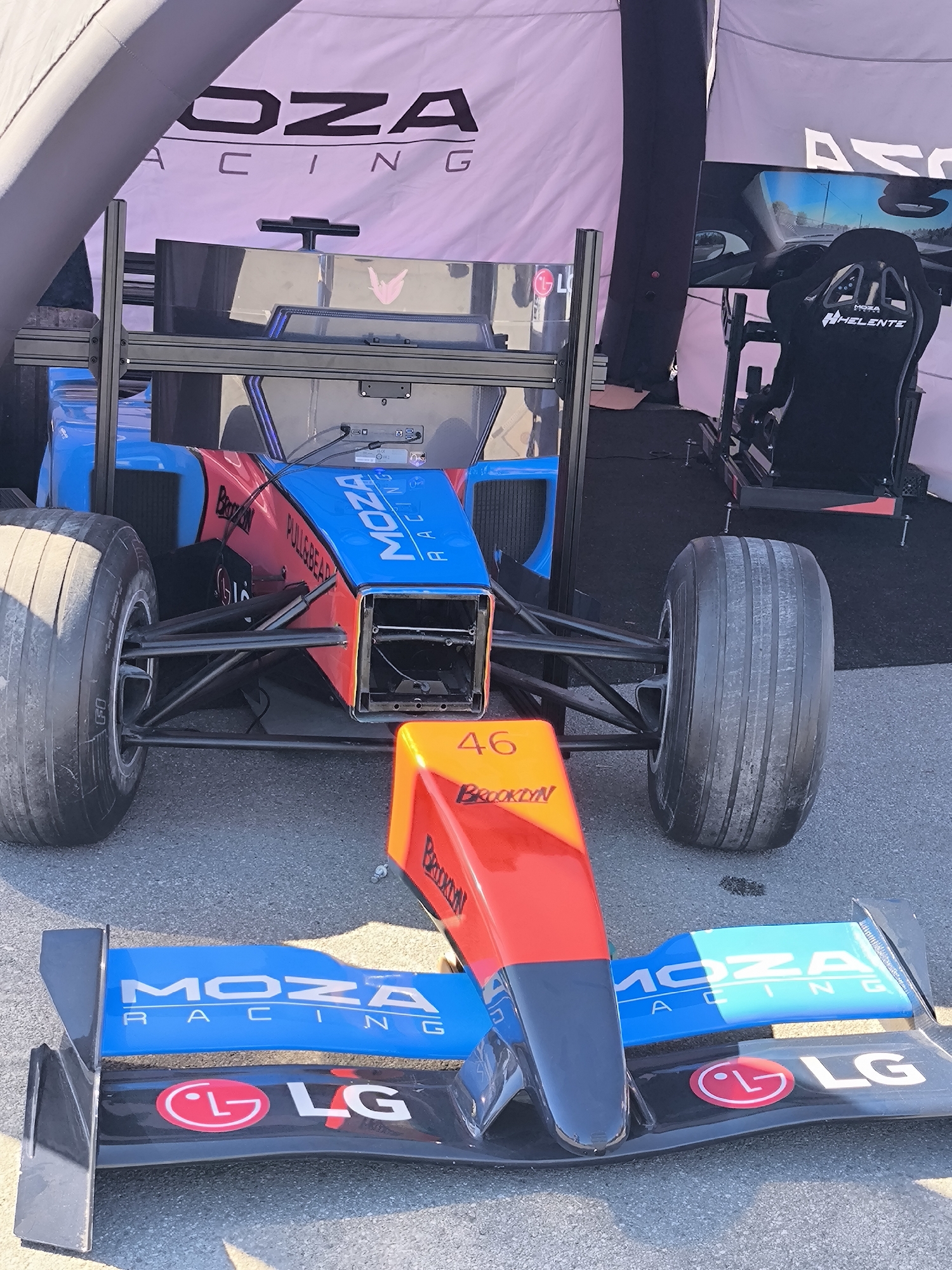 A detailed photograph shows an open-wheel race car, missing its front wing, positioned on an asphalt surface under a tent. The car's chassis is a combination of blue, red, and black, adorned with "MOZA RACING" and "BROOKLYN" branding. A front wing with similar branding and an LG logo is placed in front of the car. A racing simulator rig with a bucket seat is visible in the background. The location cannot be determined to be in Riga, Latvia based on the visual information.