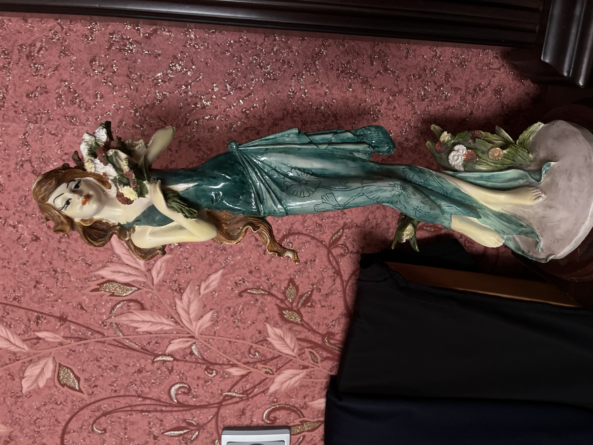 An ornate statue of a woman holding a bouquet of flowers stands in front of a textured, pink floral wallpaper. The statue depicts a woman with flowing brown hair, wearing a green dress that drapes to the floor, adorned with floral patterns. She is barefoot and stands on a circular base. To the left of the statue, a dark blue and black fabric hangs, and to the right, a dark wooden vertical beam is visible. The overall scene suggests an indoor setting, likely a home, with decorative elements. The wallpaper has a subtle shimmer, with gold glitter interspersed in the texture.