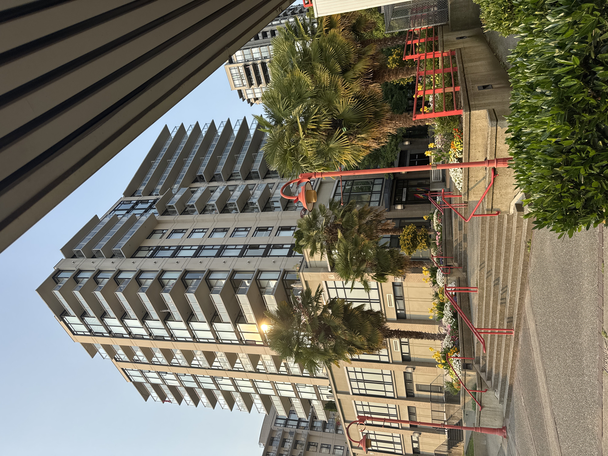 The image depicts a modern condominium building with multiple balconies, set against a clear sky. In the foreground, a concrete staircase with red railings leads up to the building's entrance. Palm trees and flower beds are strategically placed around the staircase and building, adding a touch of greenery and color. The architecture suggests an urban setting, possibly a waterfront or a well-developed residential area. The lighting indicates it's daytime, with the sun casting a warm glow on the building's facade and the surrounding vegetation. The overall scene conveys a sense of contemporary urban living with a focus on aesthetic appeal and outdoor space. The building appears to be located in North Vancouver, Canada.