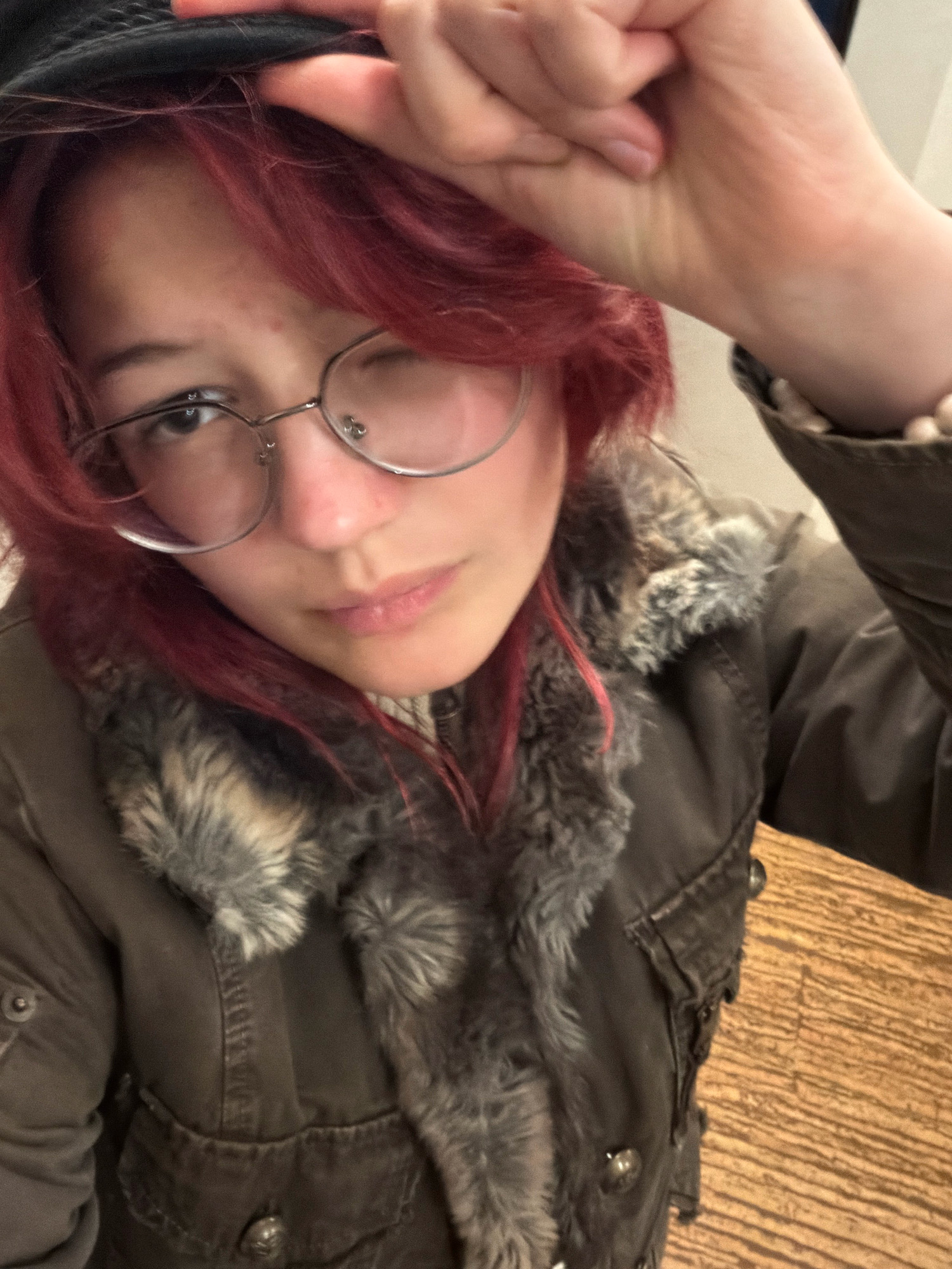 A young person with red hair and glasses is wearing a dark jacket with a fur collar and a black hat. They are holding their hand up to their head and winking. The background is a wooden floor. The image appears to be a selfie taken indoors. There is no information available to suggest this image is located in Chelyabinsk, Russia.