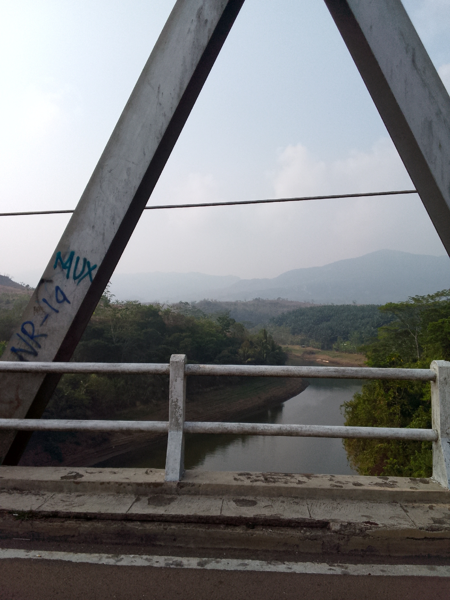 The image depicts a view from a bridge, looking out over a river and a landscape of rolling hills covered in lush vegetation. The bridge structure, made of weathered metal beams, frames the scene. Graffiti, including "MUX" and "NR-19" in blue spray paint, is visible on one of the beams. A white metal railing runs along the edge of the bridge, partially obscuring the view of the water below, which appears calm and reflects the overcast sky. The surrounding environment is predominantly green, with dense trees and palm plantations visible on the hillsides. In the distance, hazy mountains fade into the horizon. The overall atmosphere is serene and natural, suggesting a rural or semi-rural setting. The location is identified as Haurwangi, Indonesia.