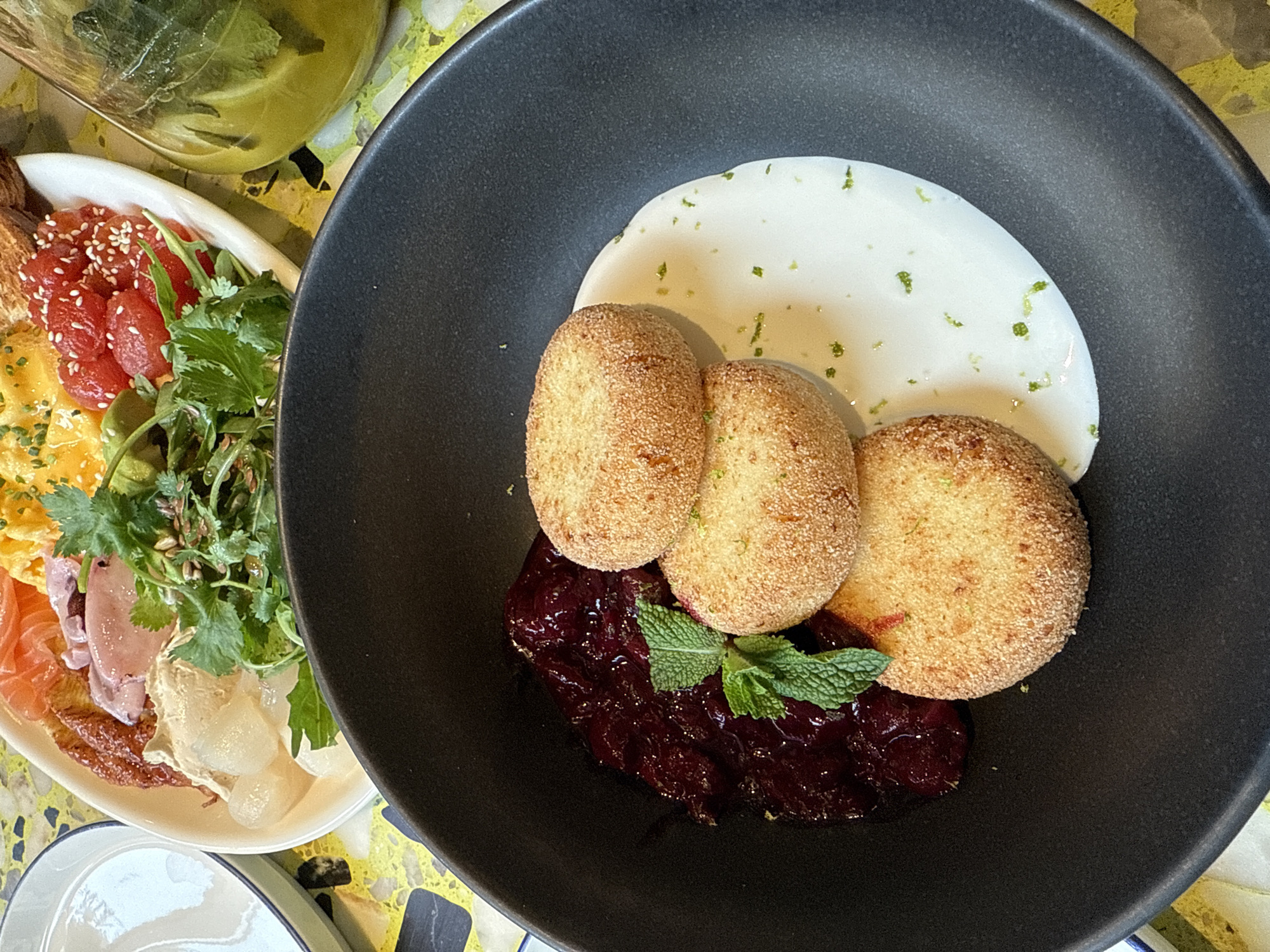 A close-up, overhead shot captures a breakfast dish in a dark bowl, placed on a yellow and white speckled table. The bowl contains three golden-brown, round patties, possibly fried cheese or croquettes, stacked slightly. To the left of the patties is a generous dollop of dark red, glistening jam or compote, garnished with a sprig of fresh mint. To the right, a smooth, white sauce, perhaps yogurt or cream, is swirled and sprinkled with fine green zest, likely lime. In the background, out of focus, a white plate holds other breakfast items, including what appears to be scrambled eggs, sliced salmon, and some diced red fruit, possibly tomatoes or berries. A clear glass jug filled with a pale green liquid and mint leaves is also visible. The scene is indoors, with the light suggesting it is daytime. There are no people or discernable city landmarks visible.