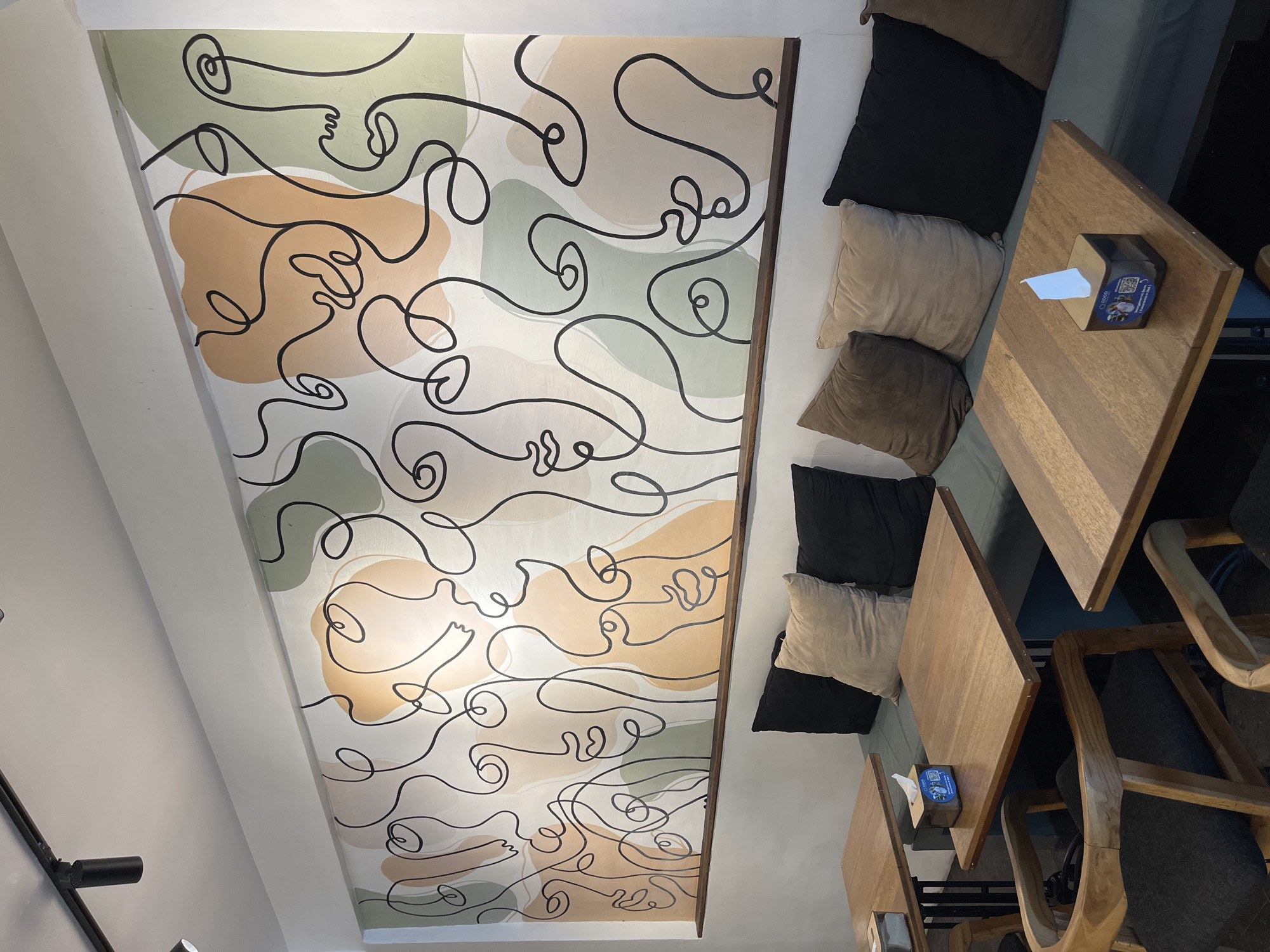 The image shows an interior space with a decorated wall, cushions, tables, and chairs, likely in a commercial setting. The wall features an abstract design of interconnected black lines over colored shapes in earthy tones of beige, green, and white.
Adjacent to the wall are cushions of varying colors (black, beige, and brown) arranged along what appears to be a bench or banquette seating area.
Two wooden tables are visible, each with a tissue box on top. Chairs with wooden frames and woven seats are positioned at the tables.
The location is Kuta Selatan, Indonesia. The specific environment suggests a cafe, restaurant, or lounge setting.