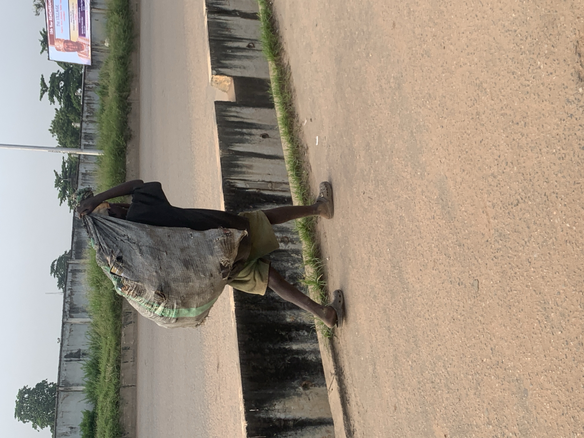 A person walks on a paved surface, carrying a large, grey, woven bag filled with contents over their left shoulder. The person is wearing a dark t-shirt, olive green shorts, and a pair of brown clogs. They are walking on a sidewalk or pathway adjacent to a road. In the background, there is a concrete wall, some overgrown green vegetation, trees, a tall light pole, and a billboard. The billboard displays a picture of a man in traditional attire and text, possibly an advertisement or public notice, which reads "10th Year REMEMBRANCE the VILLAGE". The ground is a coarse, sandy-colored asphalt. The overall scene suggests an outdoor urban environment in Shagamu, Nigeria.