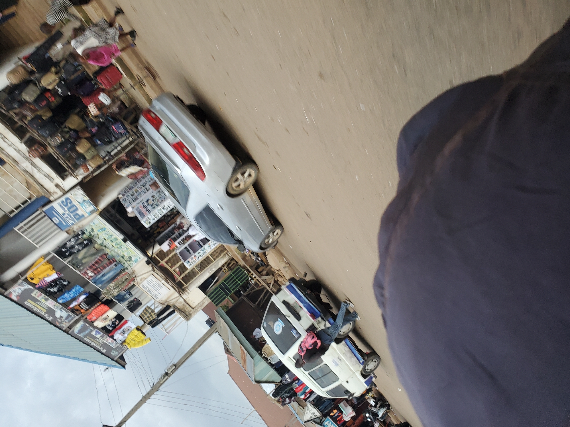 A street scene in Bukuru, Nigeria, shows a bustling market area with various shops and vehicles. In the foreground, a person's dark clothing is visible, partially obscuring the view. A white minivan with blue trim is parked on the right side of the street, and a person wearing a black jacket and blue jeans, carrying a pink backpack, is standing next to it. Further down the street, a silver sedan is parked, and several people are walking in the background. Shops lining the street display clothing and bags for sale. Power lines crisscross above the scene, and the ground is paved with dirt and gravel. The sky is overcast.