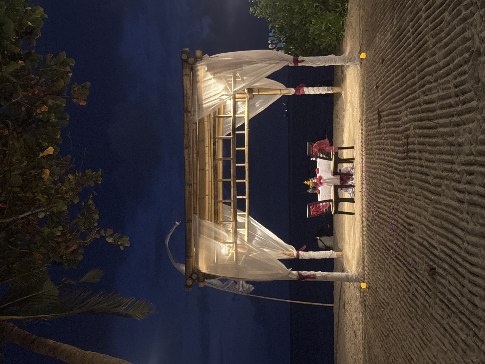 A romantic dinner setting under a bamboo structure draped with white fabric is set on a beach in Denpasar, Indonesia. The table is set for two with a white tablecloth, flowers, and what appear to be two chairs with red accents. The scene is illuminated by lighting within the structure and possibly along the path leading to it. The beach sand is textured, and faint rose petals seem to have been scattered around. The background shows a dark blue sky meeting a dark body of water, with some greenery visible to the right and left.