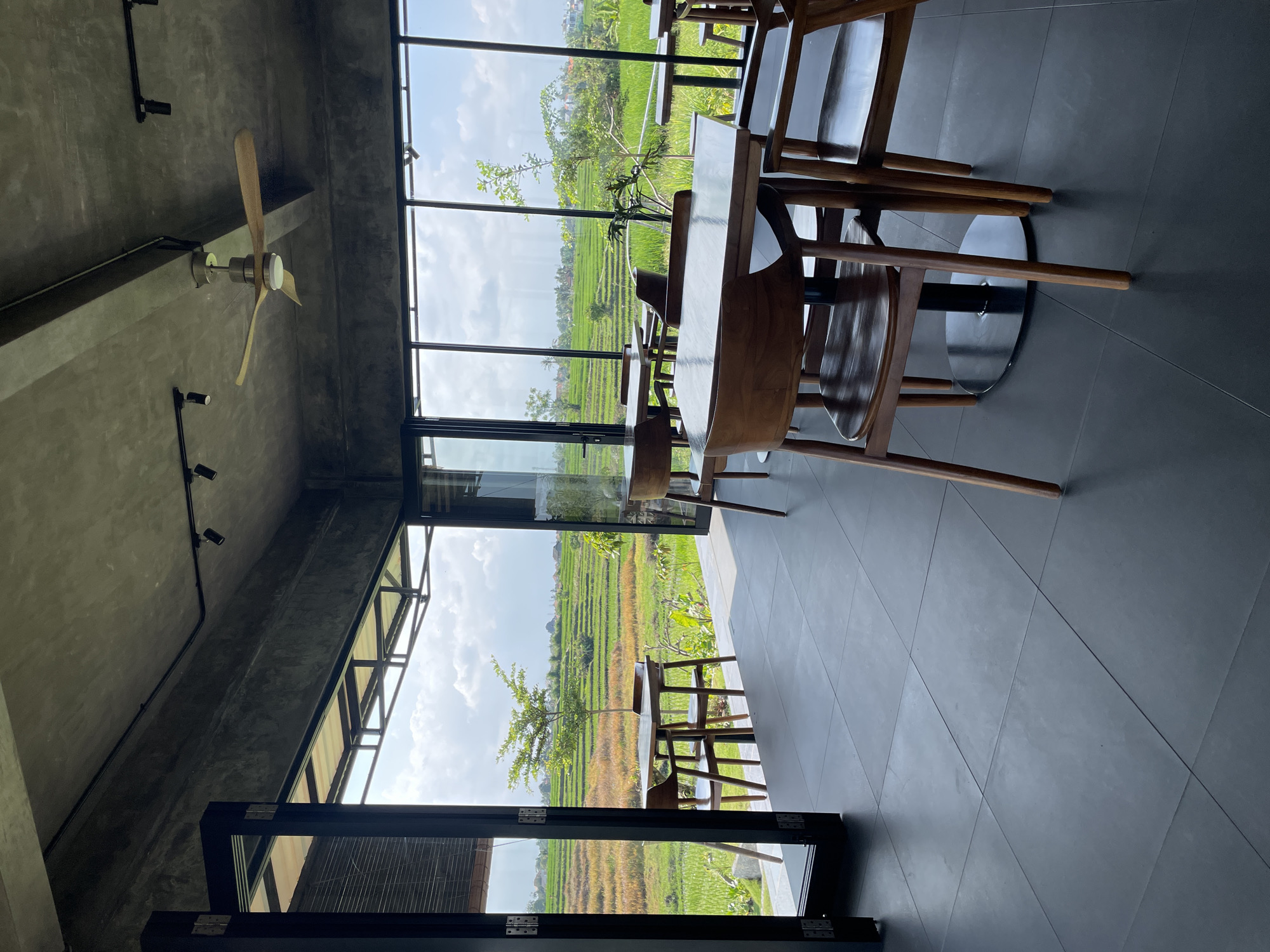 This is a medium shot, interior view of a modern cafe with large windows that overlook a lush green rice paddy field in Kediri, Indonesia. The cafe has a raw concrete ceiling with exposed track lighting and a ceiling fan. The floor is tiled with large, dark grey square tiles. Several wooden tables and chairs are arranged in the seating area. The tables are simple with dark, square tops and metal bases, and the chairs are made of dark wood with curved backs. In the foreground, a table with chairs is positioned to the right, and further back, another table with chairs is visible. Through the large windows, the expansive terraced rice fields stretch out, dotted with a few trees and small buildings in the distance. The sky is partly cloudy and bright. No people are visible in the frame. The overall atmosphere is serene and airy, emphasizing the connection between the indoor space and the natural landscape.